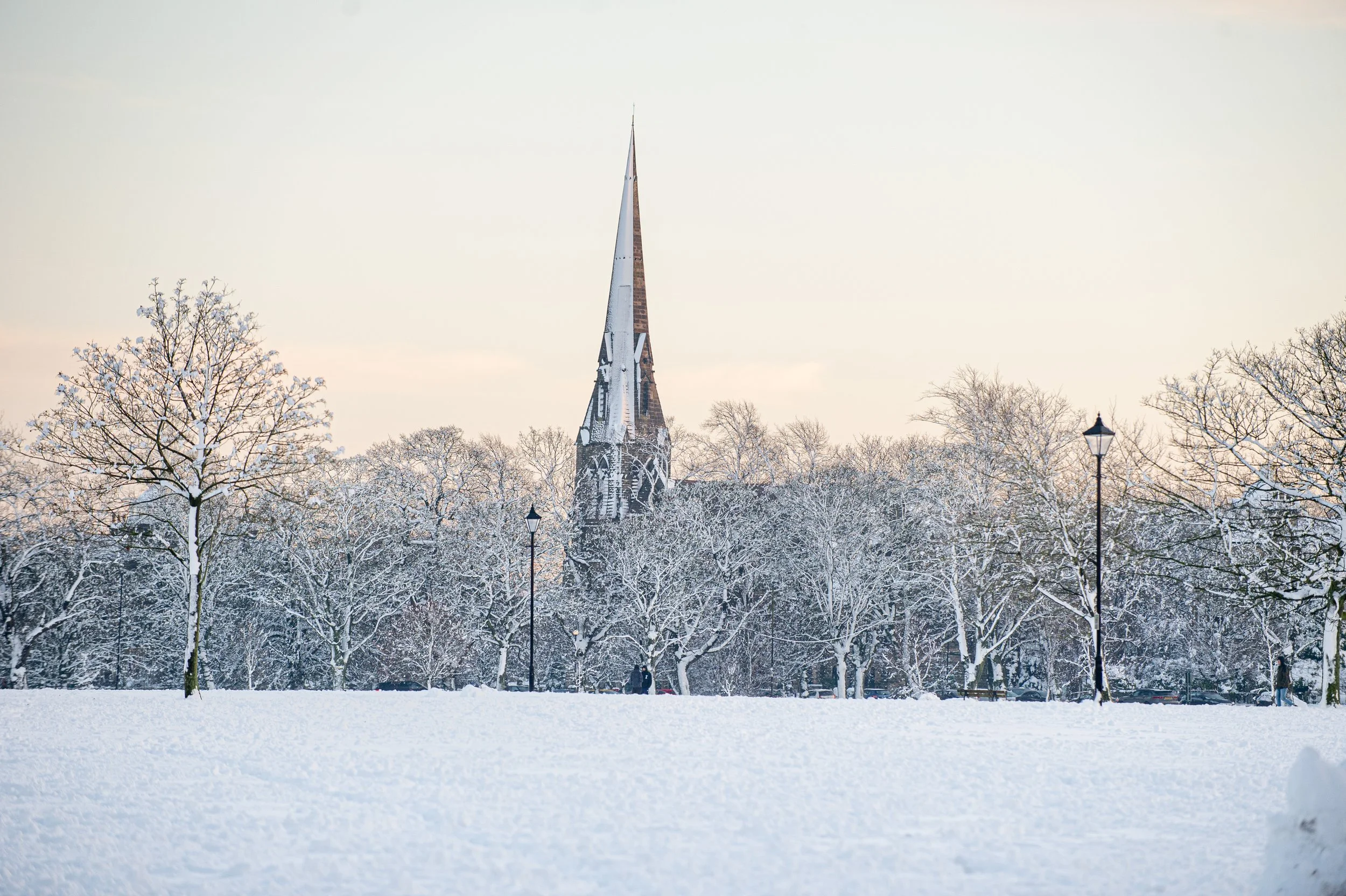 Snow-covered park with leafless trees, street lamps, and a church with a tall steeple in the background.