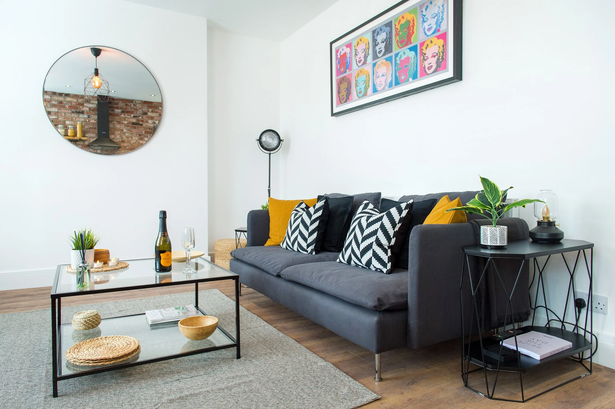 Living room with white walls, a dark gray sofa with black and gold pillows, a glass coffee table with decorative items, a brick wall reflected in a round mirror, colorful pop art portrait of Marilyn Monroe on the wall, a potted plant, and a black sid