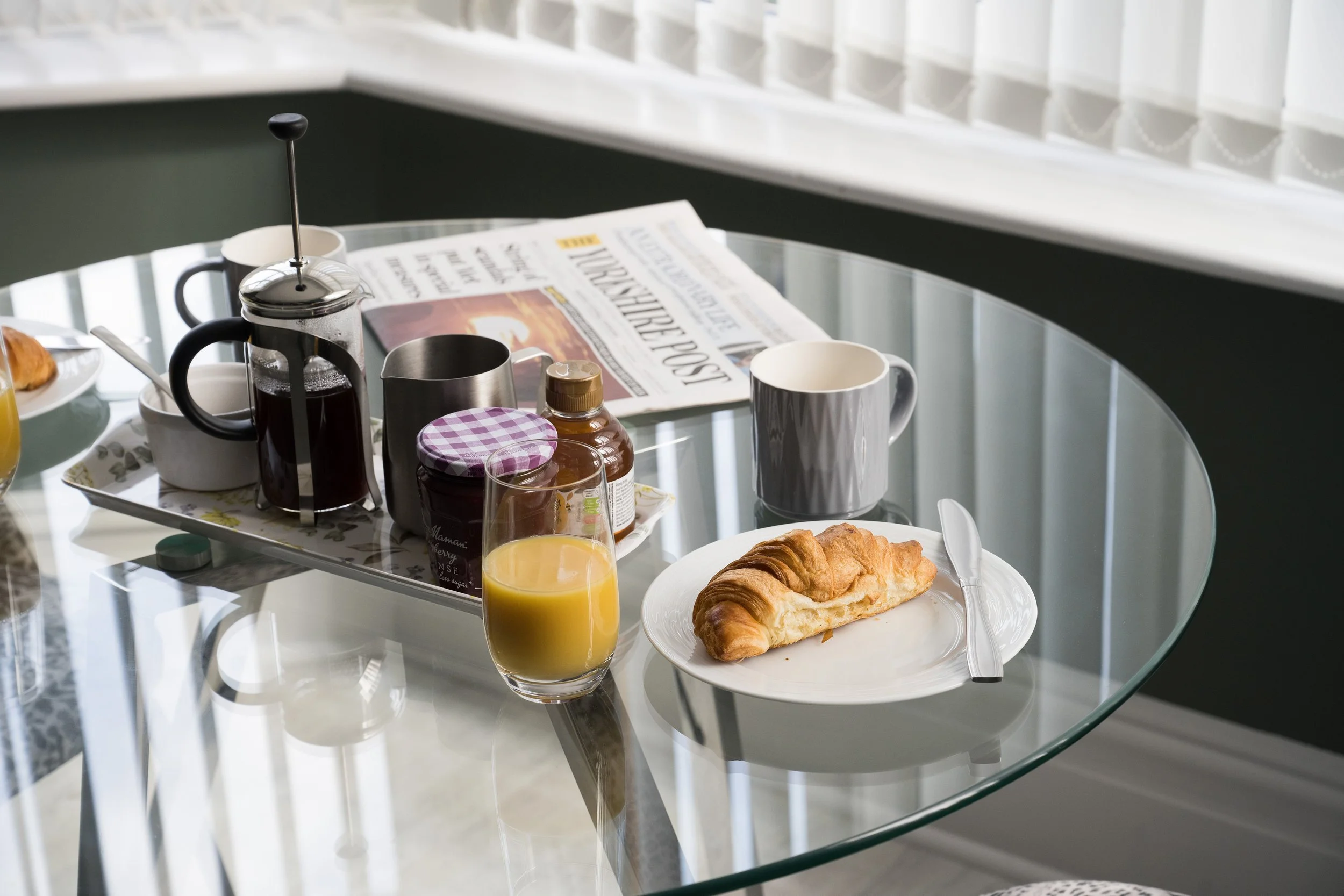 A breakfast table with a croissant on a white plate, a glass of orange juice, a cup, a French press coffee maker, a jar of jam, a newspaper, and a second mug on a glass top table in a bright room with a window.