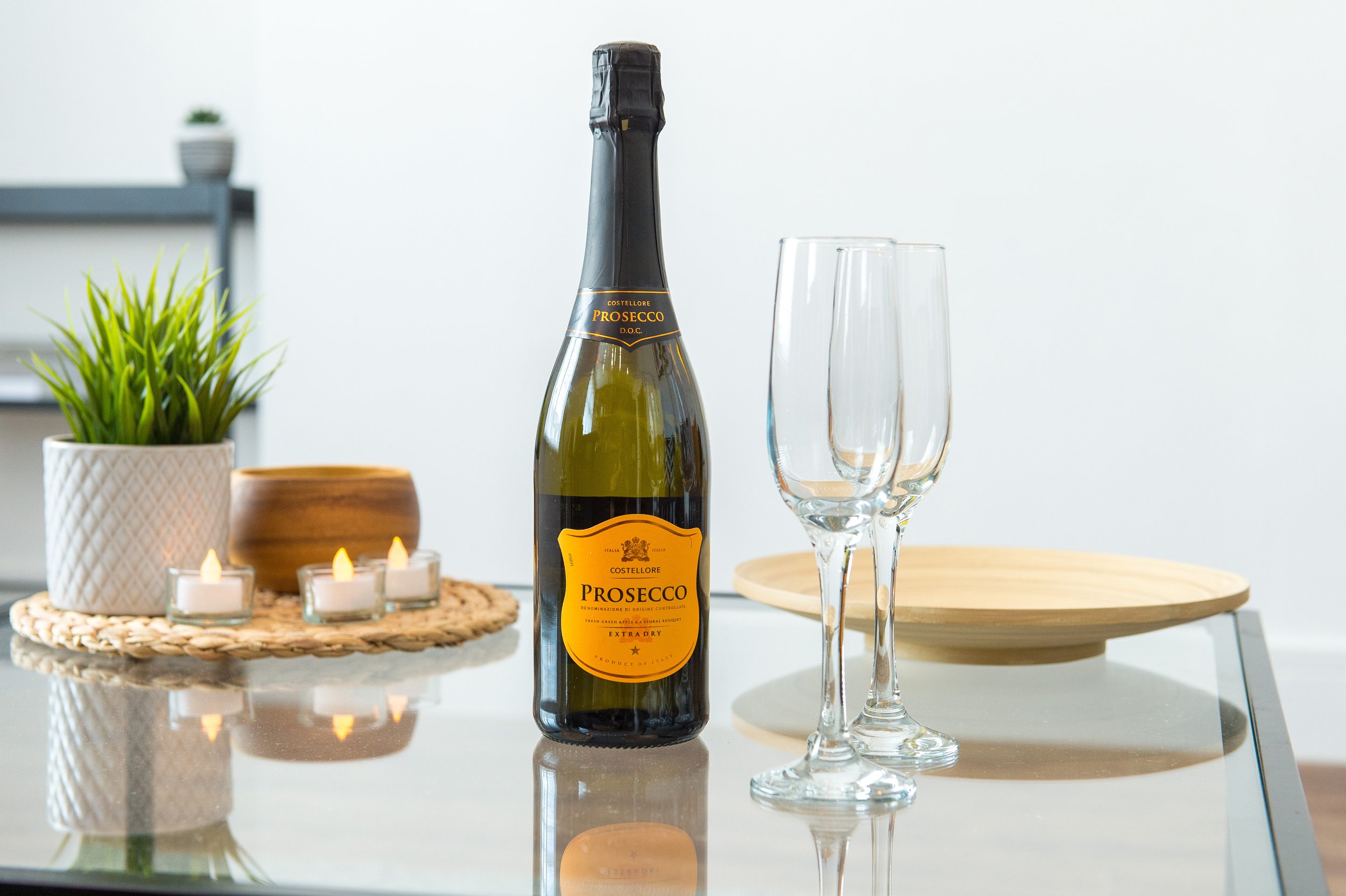 A bottle of Prosecco on a glass table with two empty champagne glasses, a small potted plant, a wooden bowl, and tea light candles for a celebration.