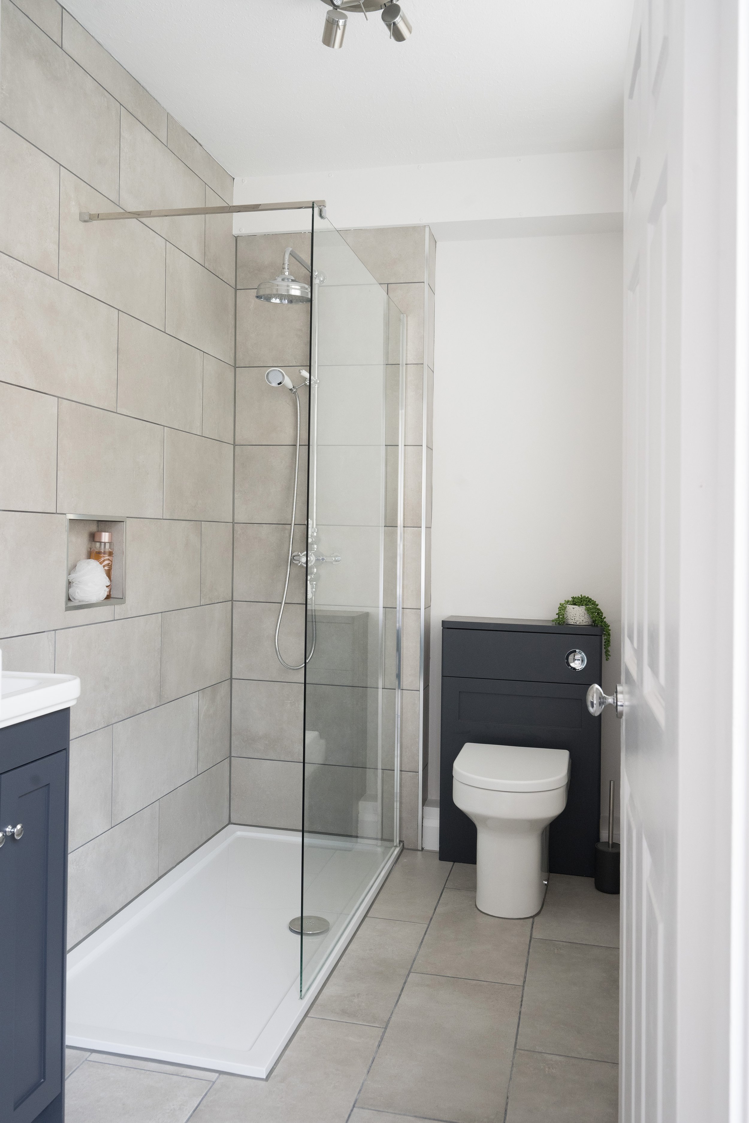 A modern bathroom featuring a walk-in shower with a glass partition, beige tiled walls, a white toilet, and a dark gray cabinet with a potted plant on top. The bathroom has a white ceiling with a ceiling light fixture and light-colored floor tiles.