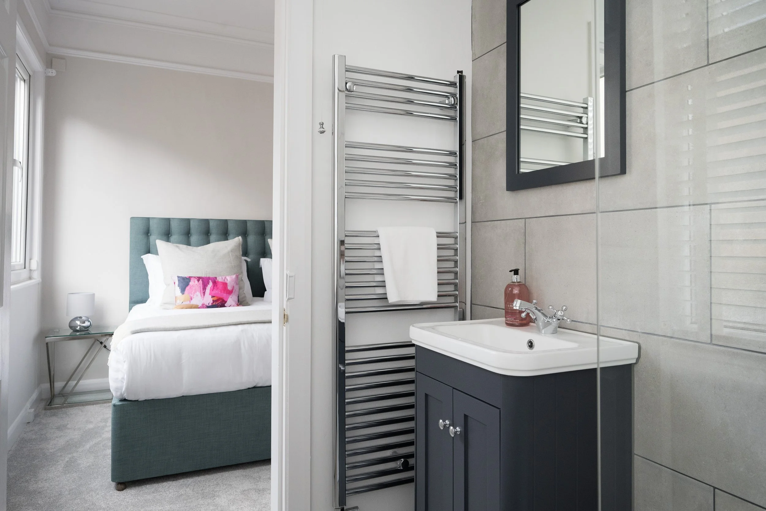 A bedroom visible through an open doorway, with a bed that has a teal upholstered headboard and a colorful pillow, a bedside table with a silver lamp, and a window. To the right, a bathroom with a gray tiled wall, a mirror with a black frame, a sink 