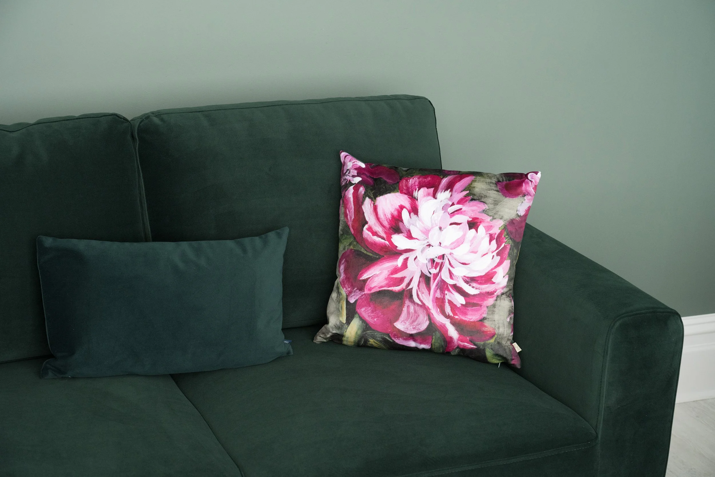 A green sofa with three cushions, one dark green, one matching the sofa, and a vibrant pink and white floral cushion, against a plain light-colored wall.