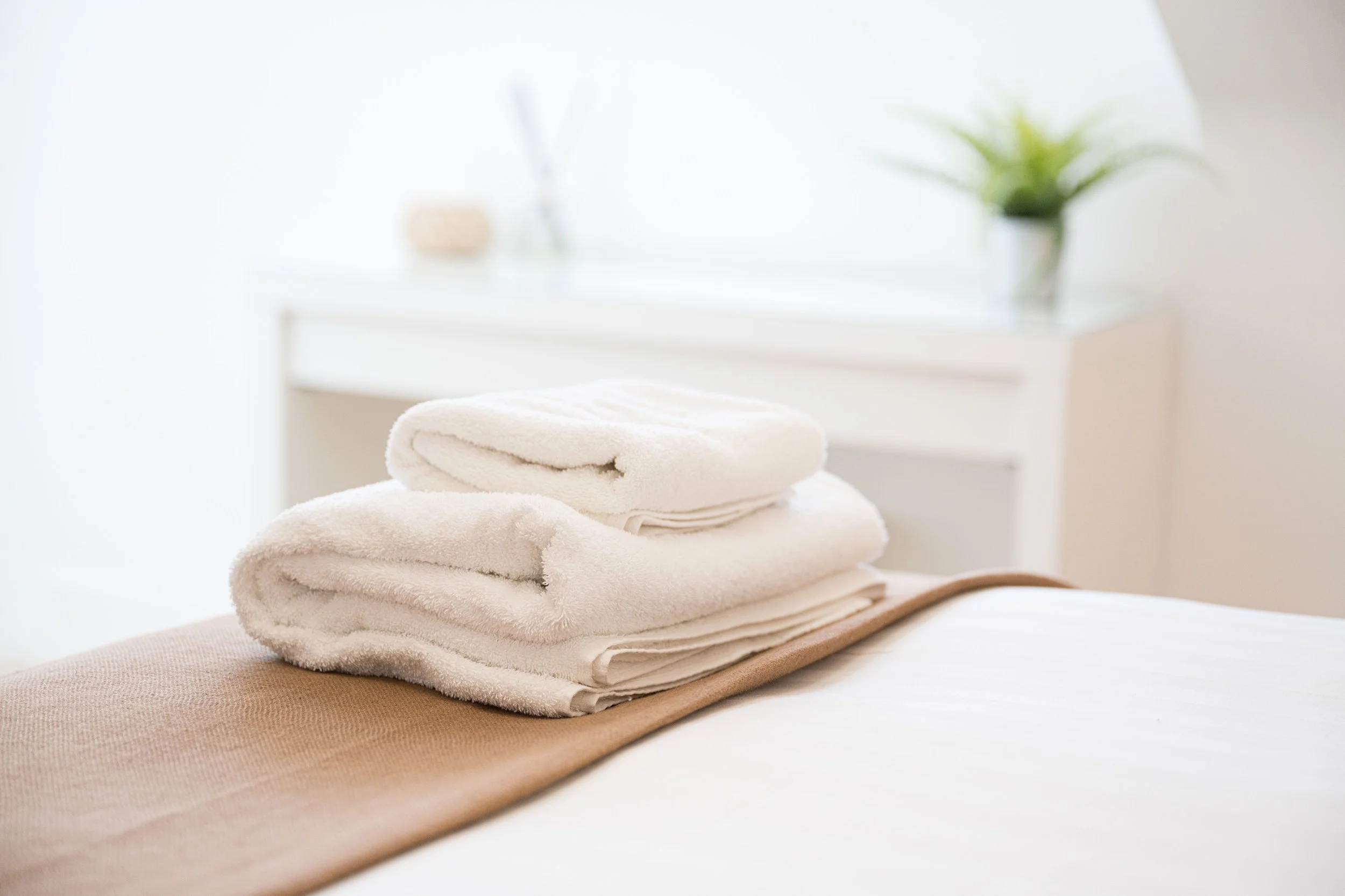 Stacked white towels on a bed with beige sheet in a bright, minimalist room with a white table and potted plants in the background.