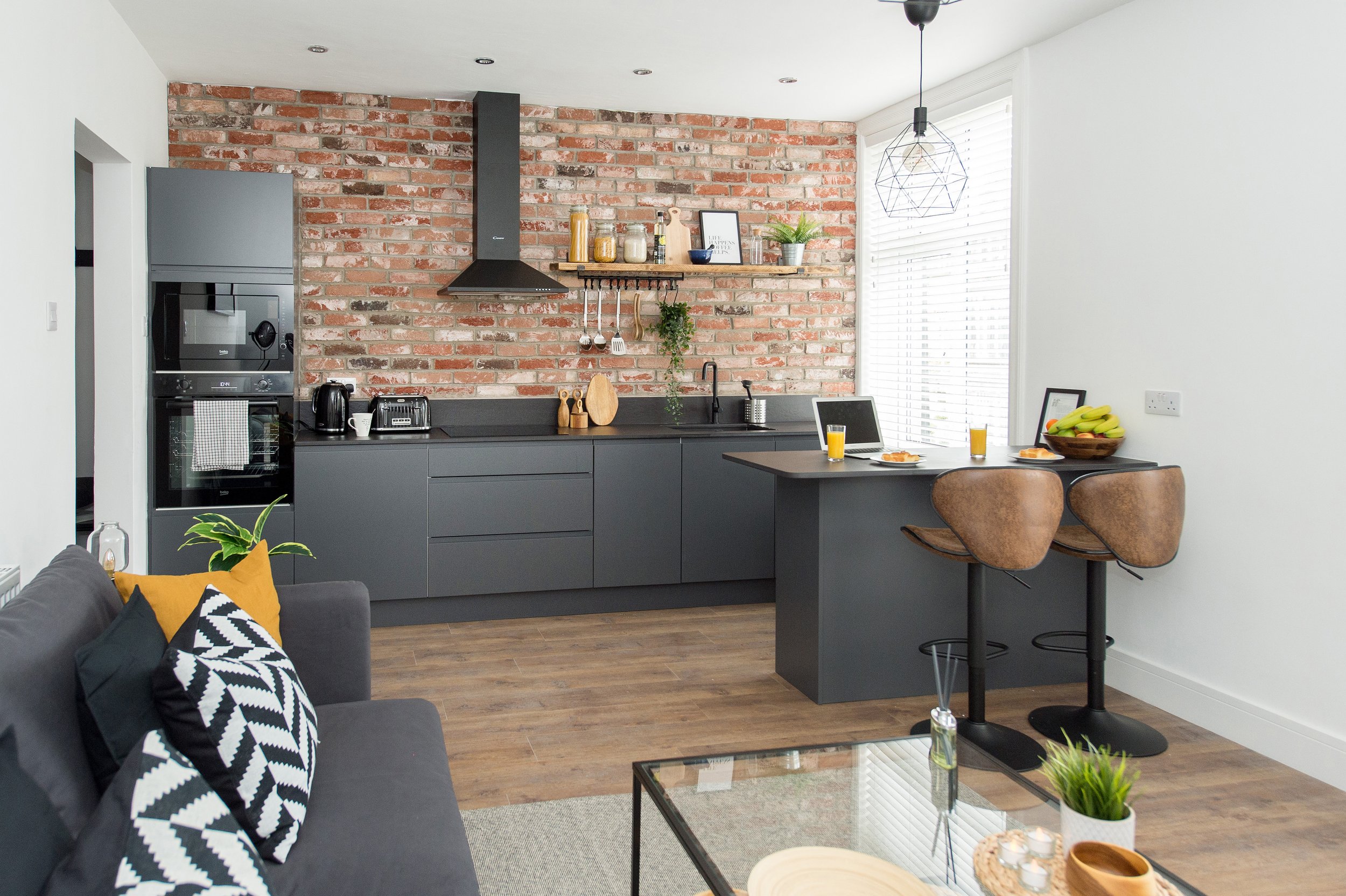 Modern kitchen with brick backsplash, gray cabinetry, open floating shelf with jars and decor, black kitchen countertop with sink, breakfast bar with two brown leather chairs, large window with white blinds, pendant light fixture, part of living room
