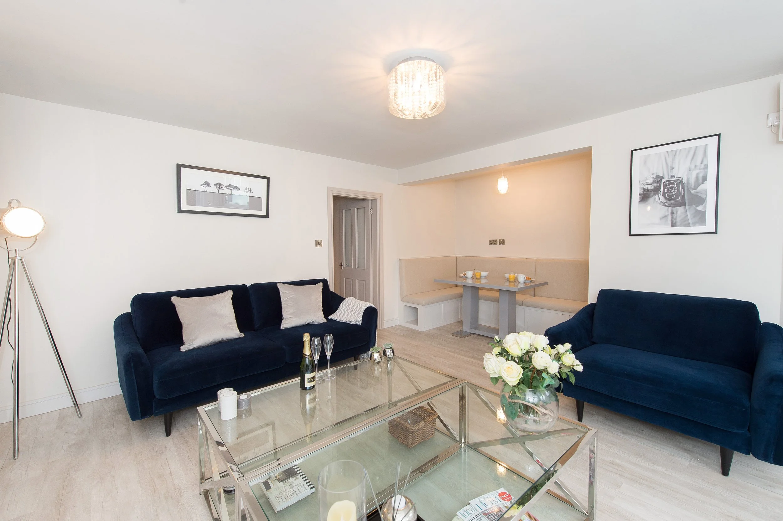 A living room with white walls, a ceiling light fixture, black framed art, navy sofas with pillows, a glass coffee table with candles, drinks, a flower arrangement, and a dining nook with a table and built-in seating.