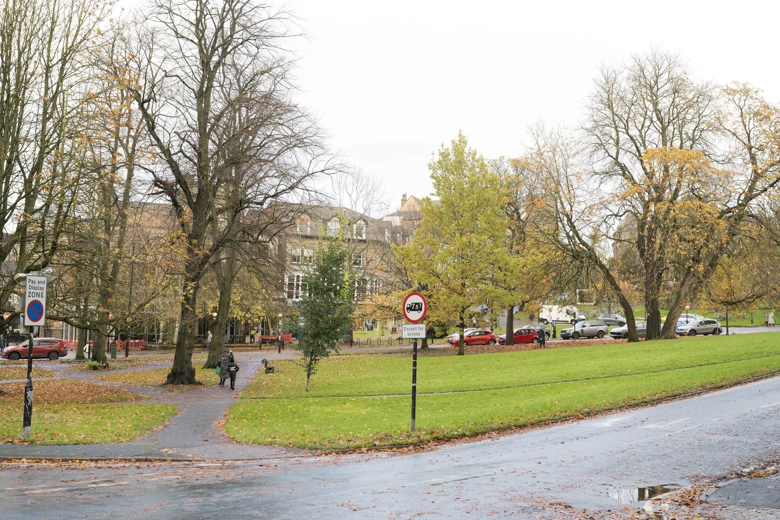 A park scene with leafless and some green trees, a walking path, parked cars, and a few people walking. There are multiple signs in the area, including a no parking zone sign and a truck limit sign.