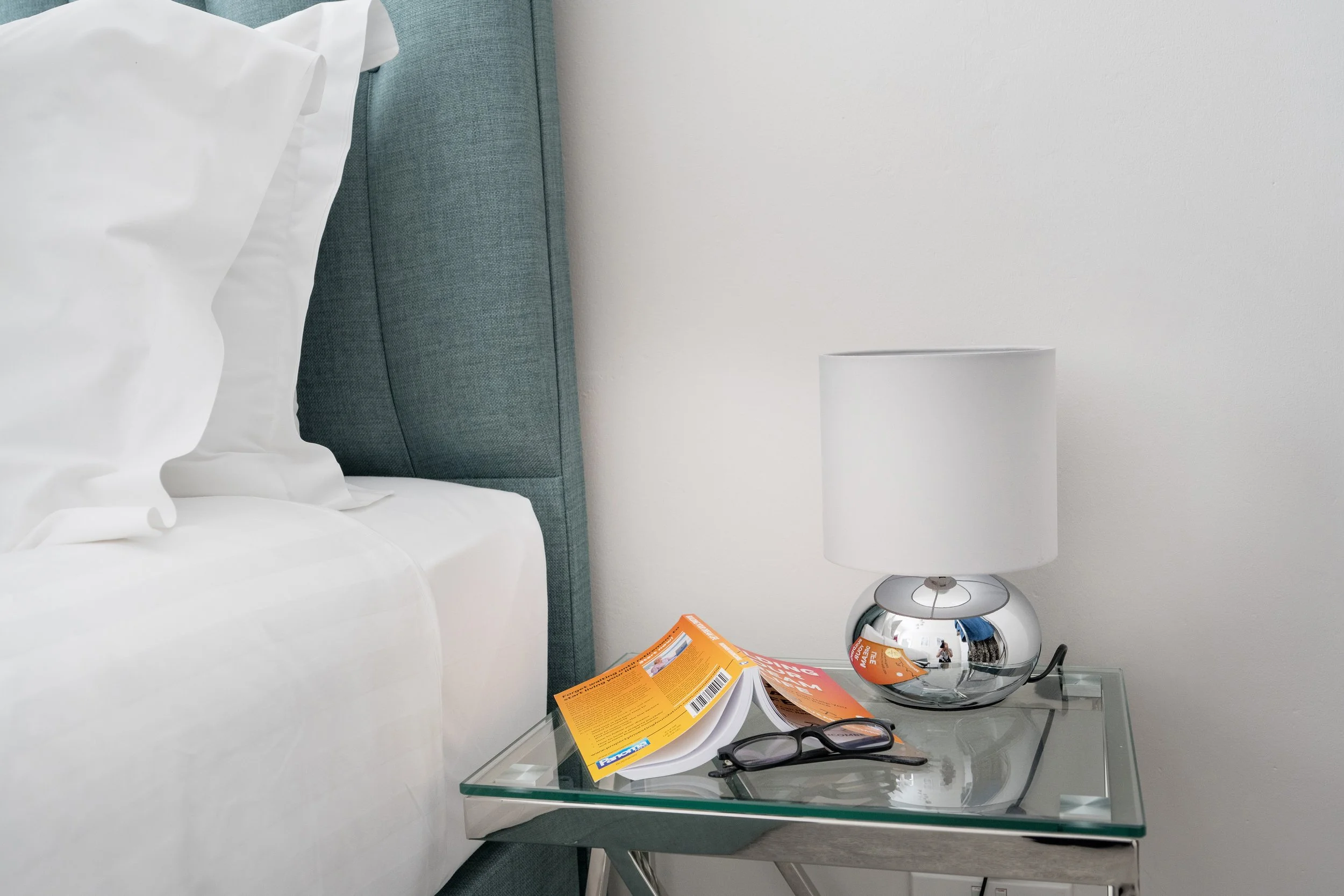 Close-up of a bedside table with magazines, eyeglasses, and a lamp in a bedroom with a white bed and teal headboard.