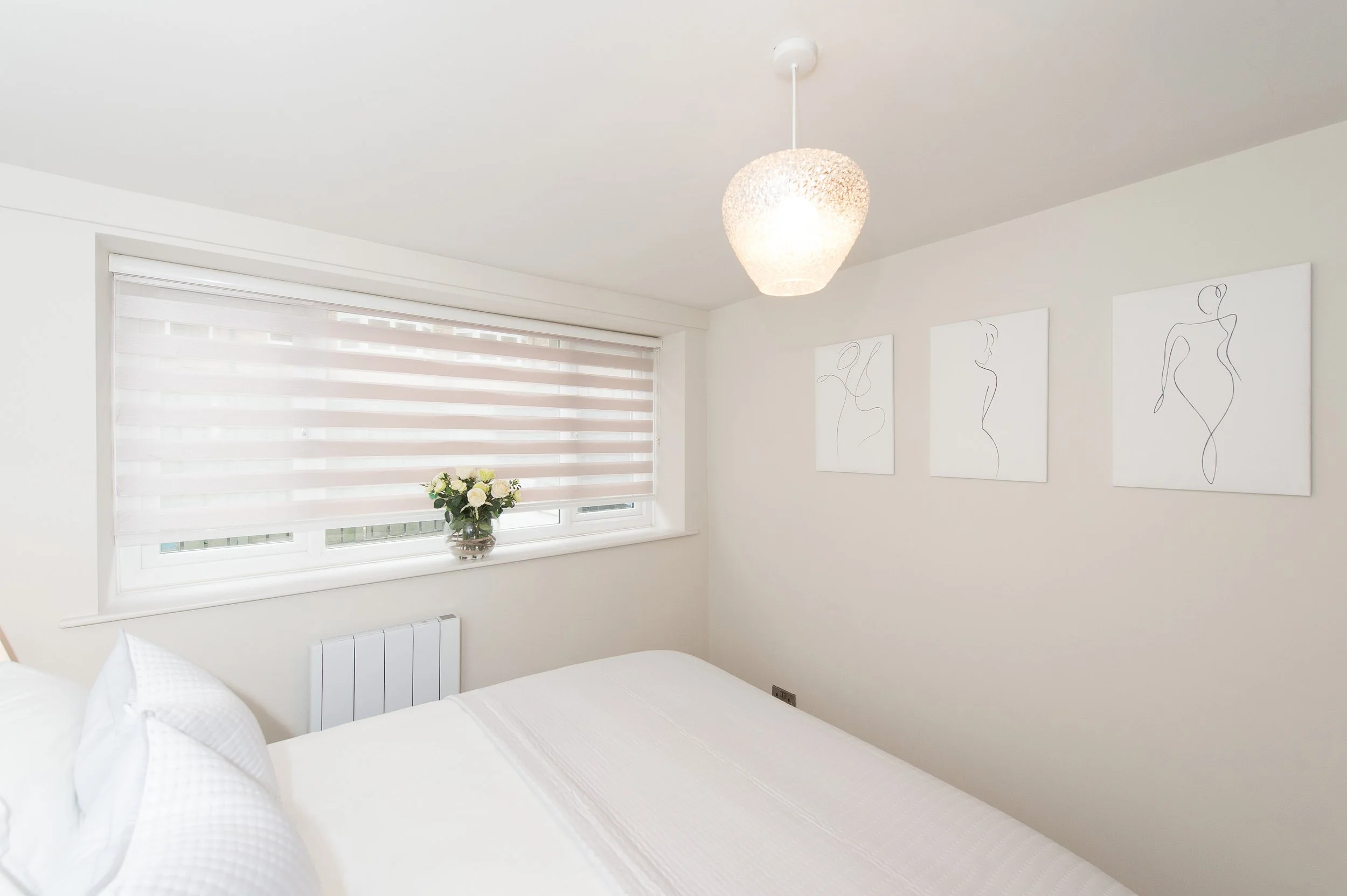 A white bedroom with a bed, a window with horizontal blinds, a vase of white flowers on the windowsill, three minimalist line art portraits on the wall, and a ceiling light.