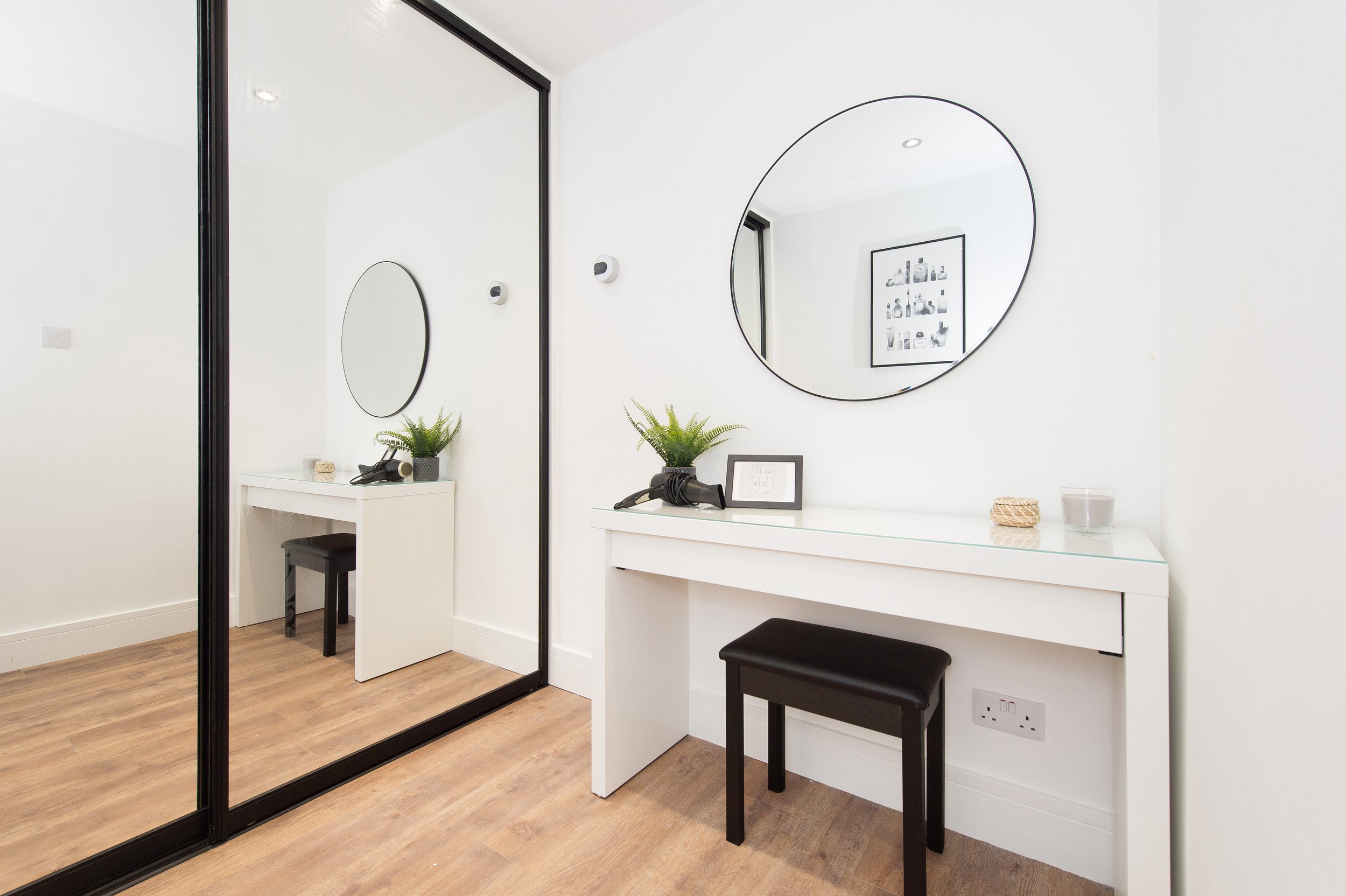 Interior view of a modern dressing area with white walls, two large round mirrors, black-framed glass sliding doors, a white vanity table with plants and accessories, a black bench, and a framed black-and-white art piece on the wall.