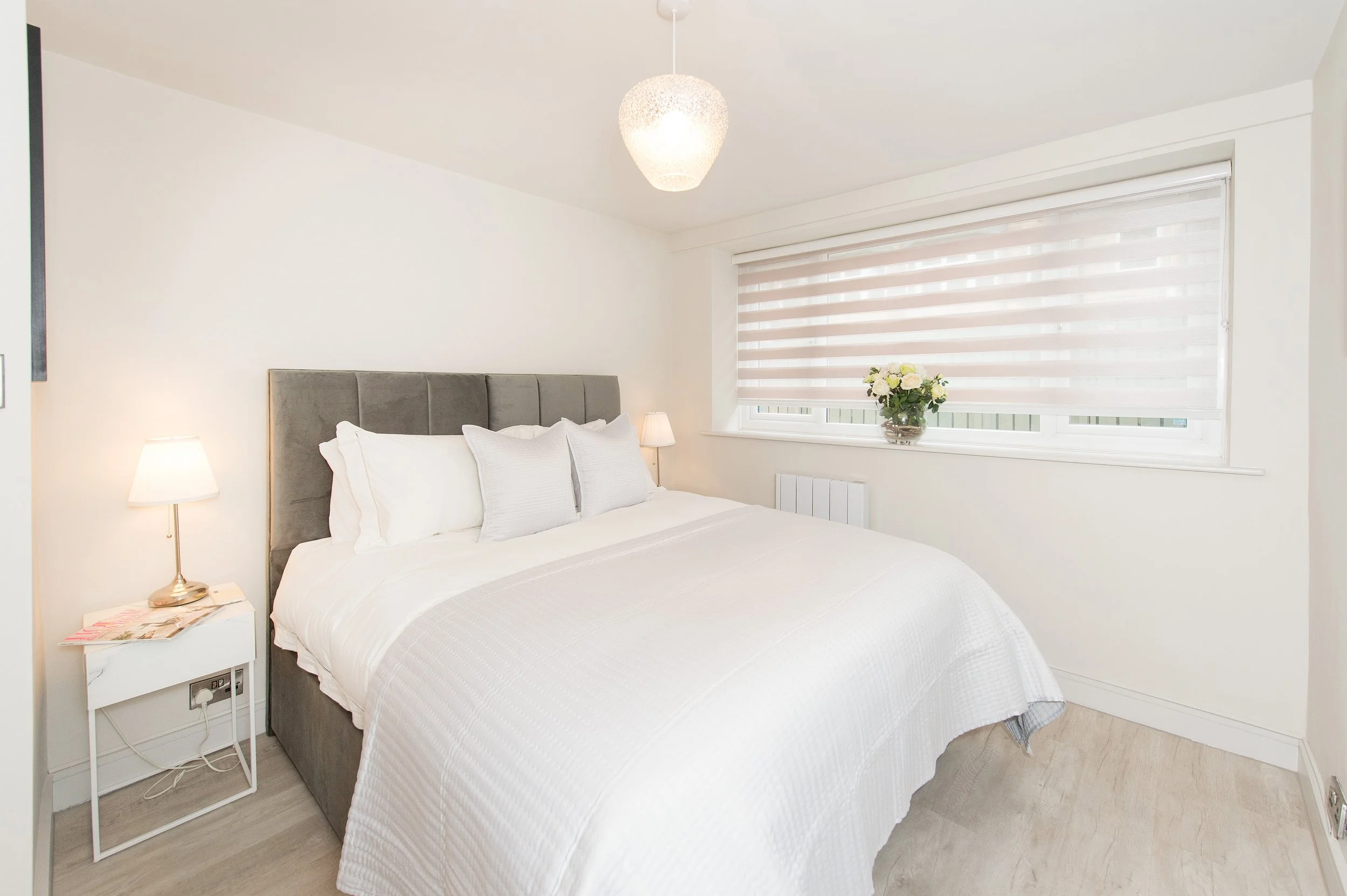 A bedroom with a bed, white bedding, a gray headboard, small white nightstand with a lamp and magazines, a window with blinds and a flower vase, and a ceiling light fixture.