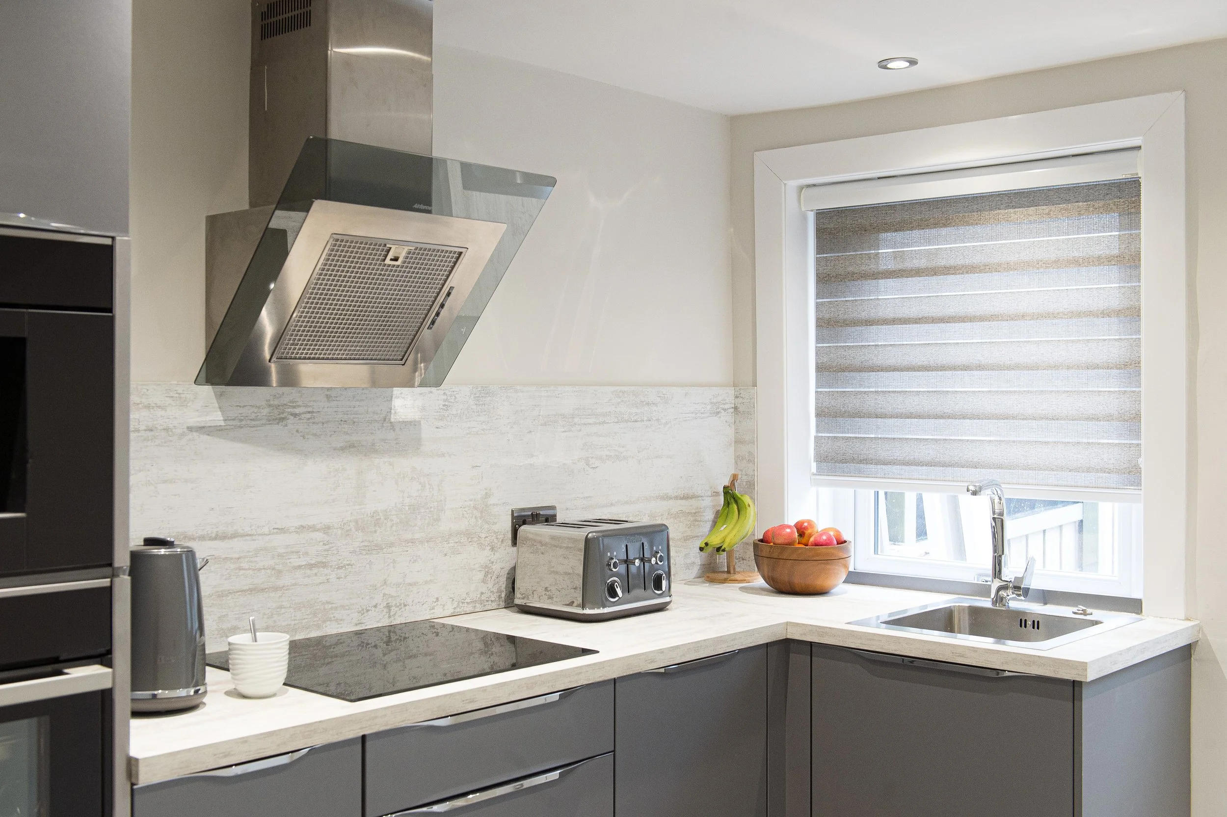 Modern kitchen with white countertop, grey cabinets, stainless steel range hood, toaster, bananas, apples, and a bowl on the counter, window with beige striped blinds.