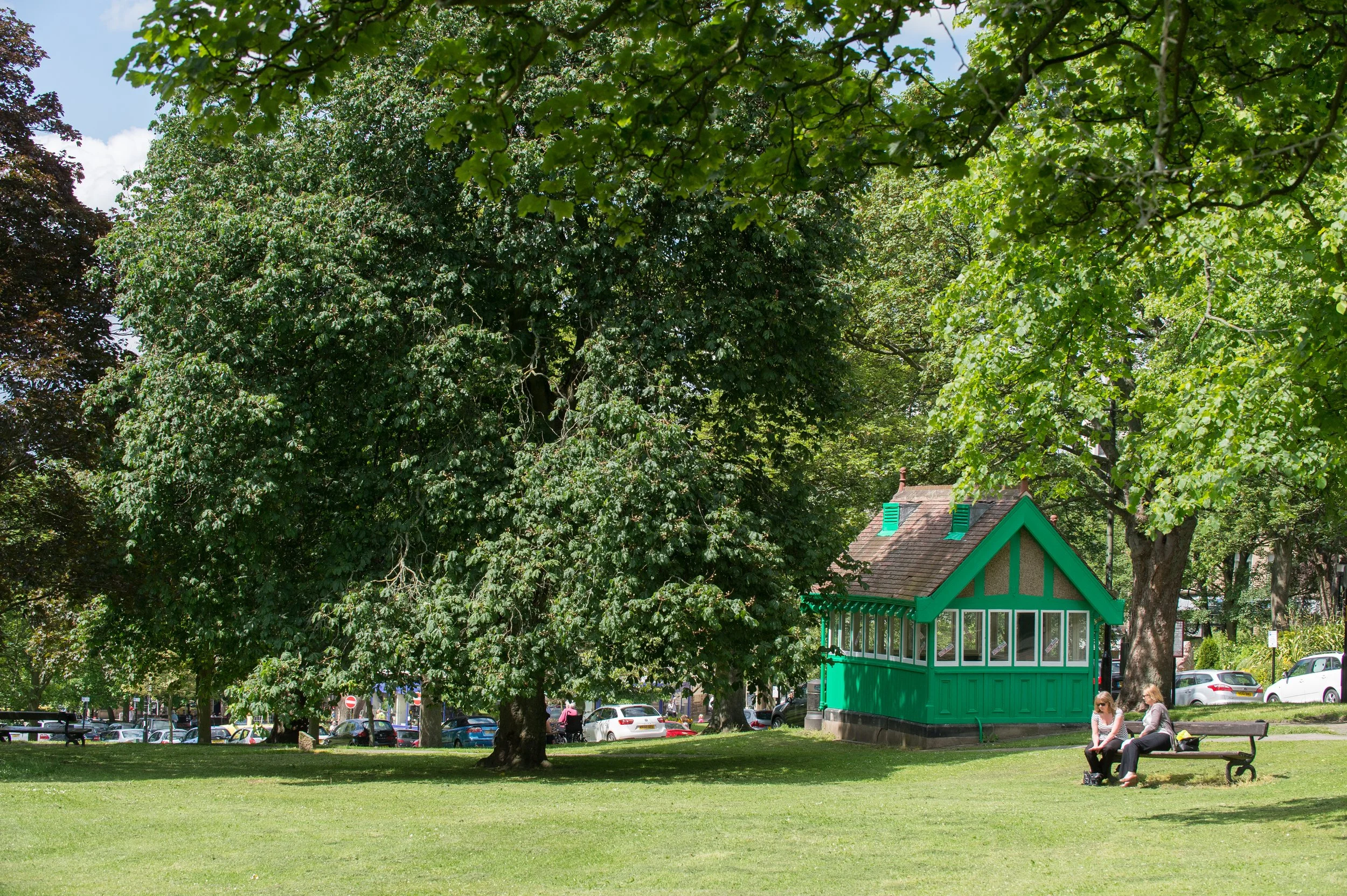 A park scene with large leafy trees, a small green building with windows, a grassy area with two women sitting on a bench, and parked cars in the background.