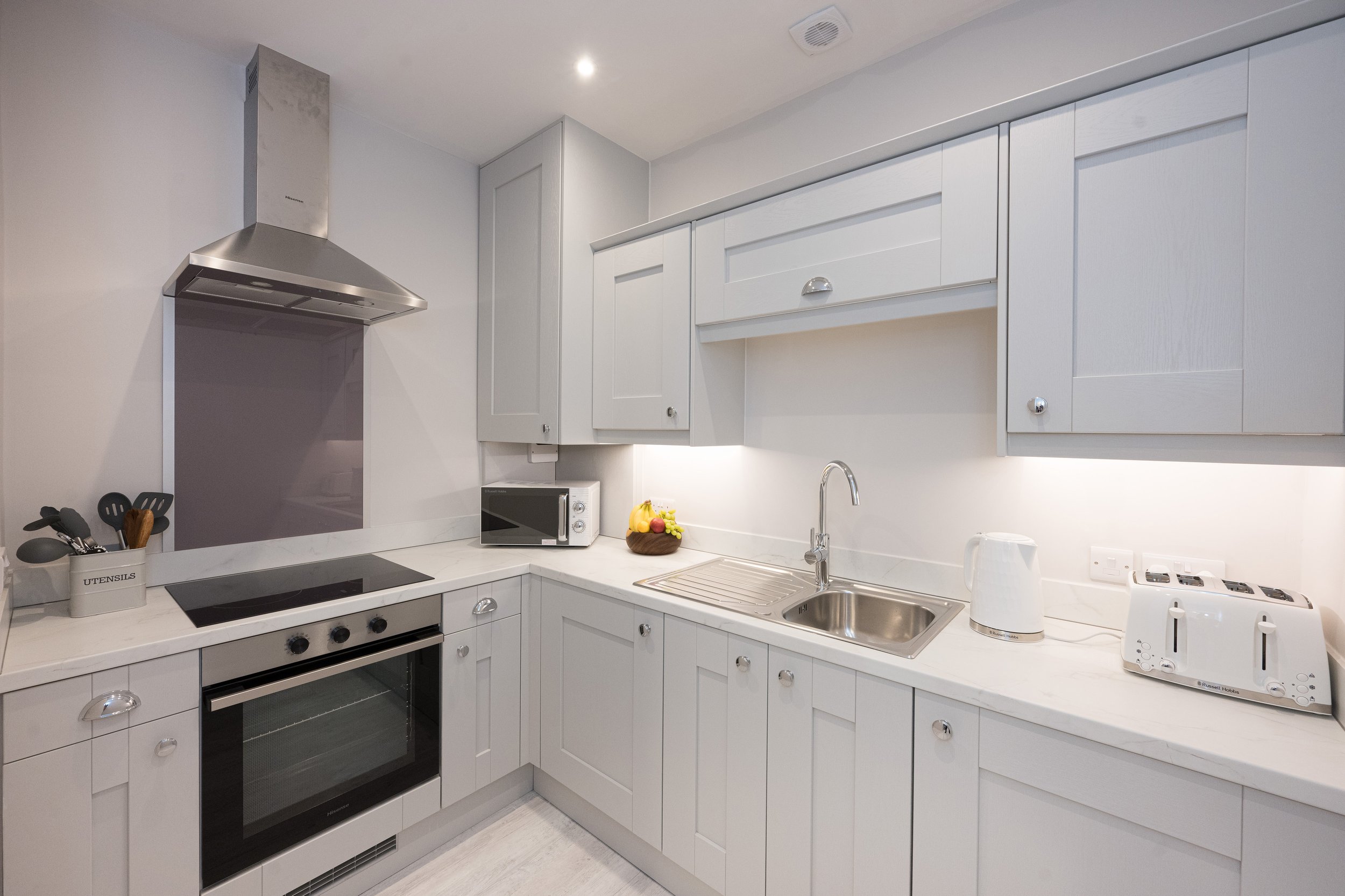 A modern white kitchen with a stove, microwave, sink, and countertop appliances including a toaster and kettle. White cabinets and drawers with round knobs, a fruit bowl, and utensils in a container labeled 'UTENSILS' are visible.