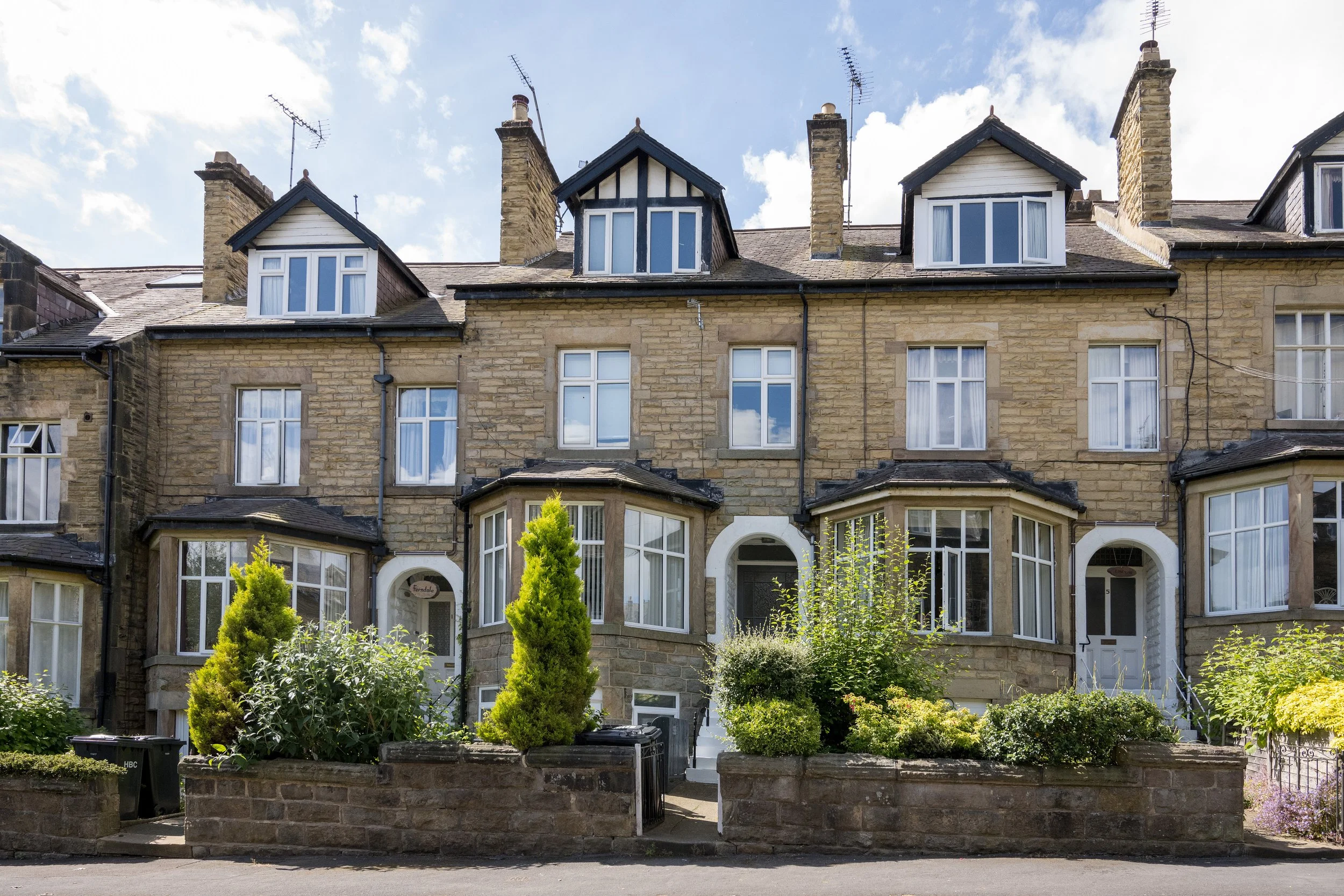 A row of tan stone townhouses with white-framed windows and dark roofs, featuring three prominent mansard-style dormer windows on the second floor. There is a small garden with greenery and a brick wall in front, along with a street in the foreground
