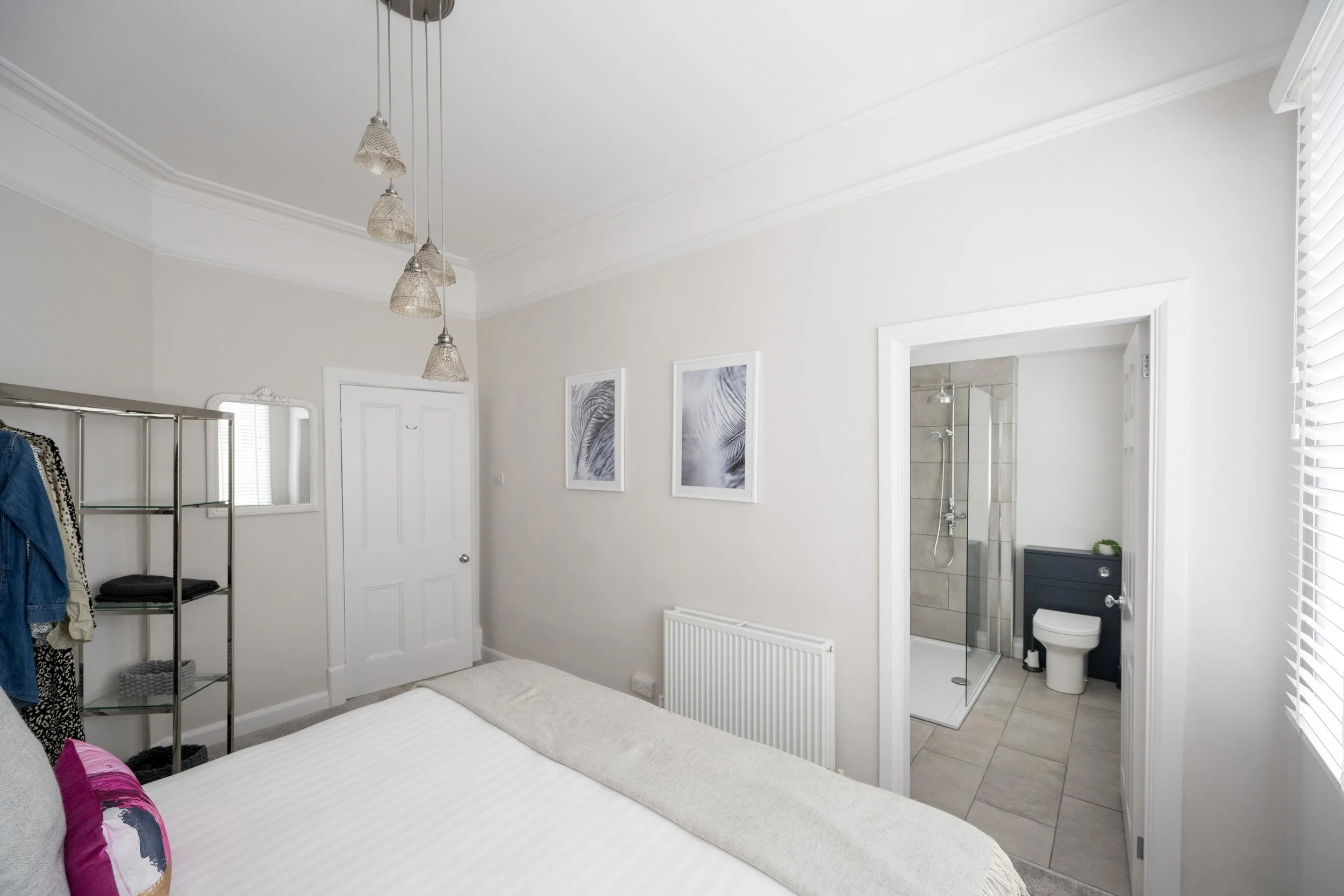 A bedroom with a white bed, purple and white pillows, and a light-colored rug. Wall art includes black and white photos of leaves. An open door reveals a bathroom with a walk-in shower and toilet. A window with white blinds is on the right. There's a