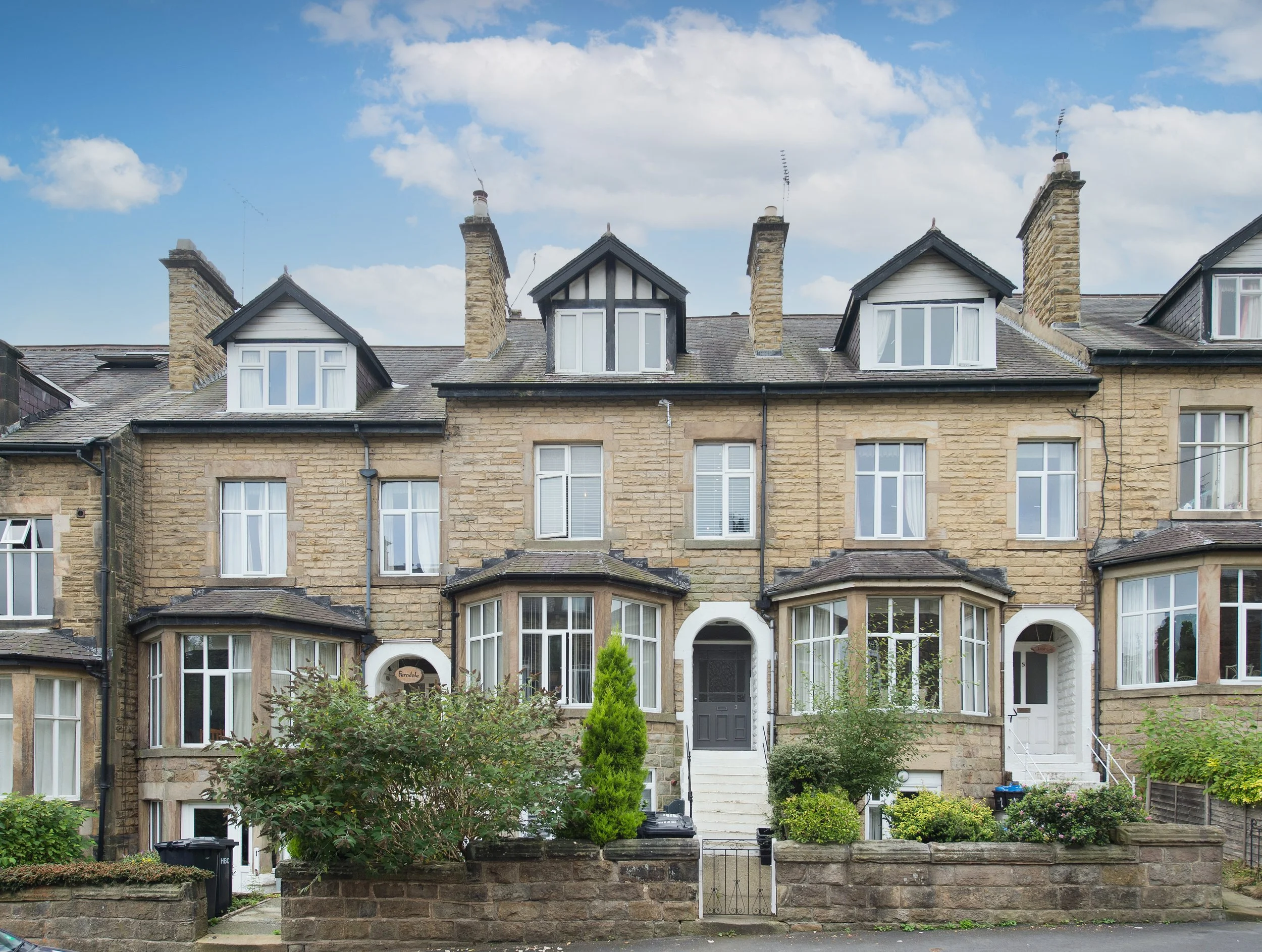 A row of old brick townhouses with dormer windows on the roof, small front yards with bushes and plants, and concrete steps leading to the doors. The sky is partly cloudy.