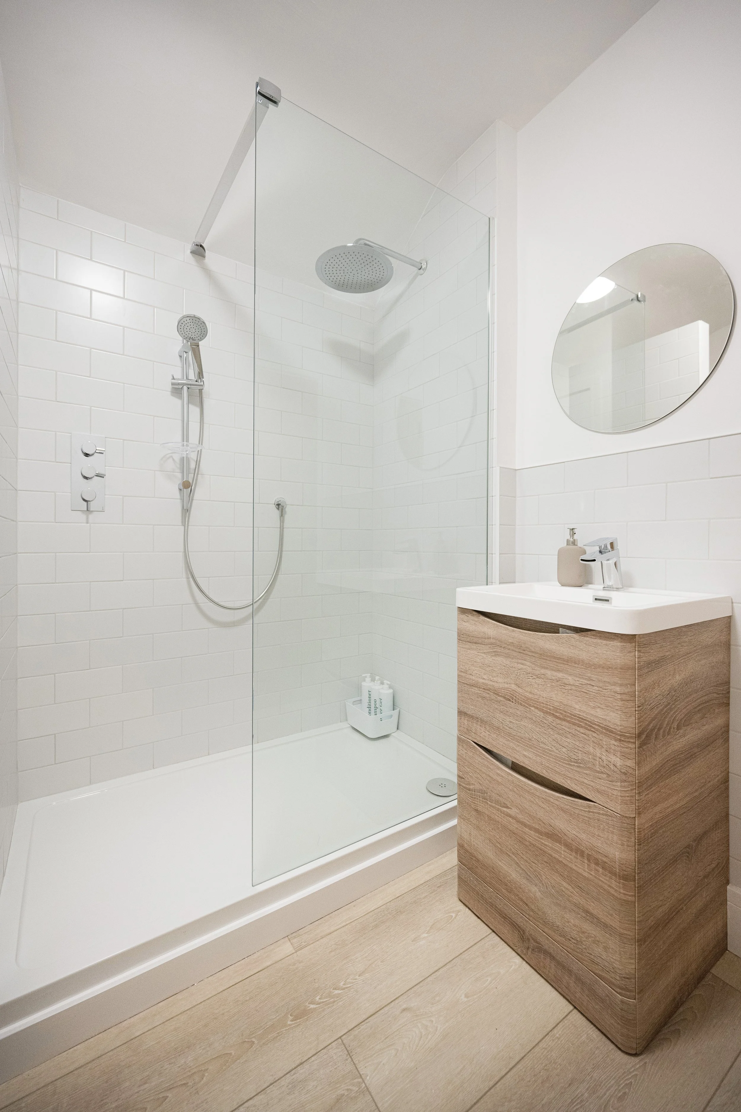 Modern bathroom with walk-in shower, glass partition, white subway tile walls, and a wooden vanity with a small sink and mirror.