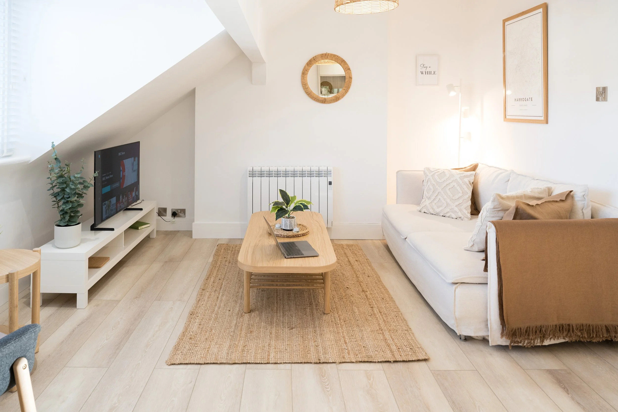 Bright living room with white walls, a cream sofa with cushions, a wooden coffee table with a laptop, a flat-screen TV on a white stand, potted plants, framed artwork on the wall, and a beige rug.
