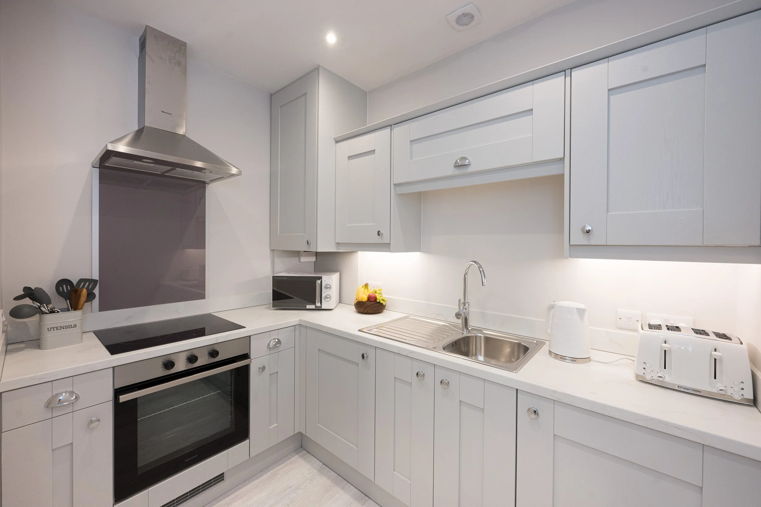 A modern white kitchen with cabinets, a stove, microwave, sink, and countertop with a bowl of fruit.