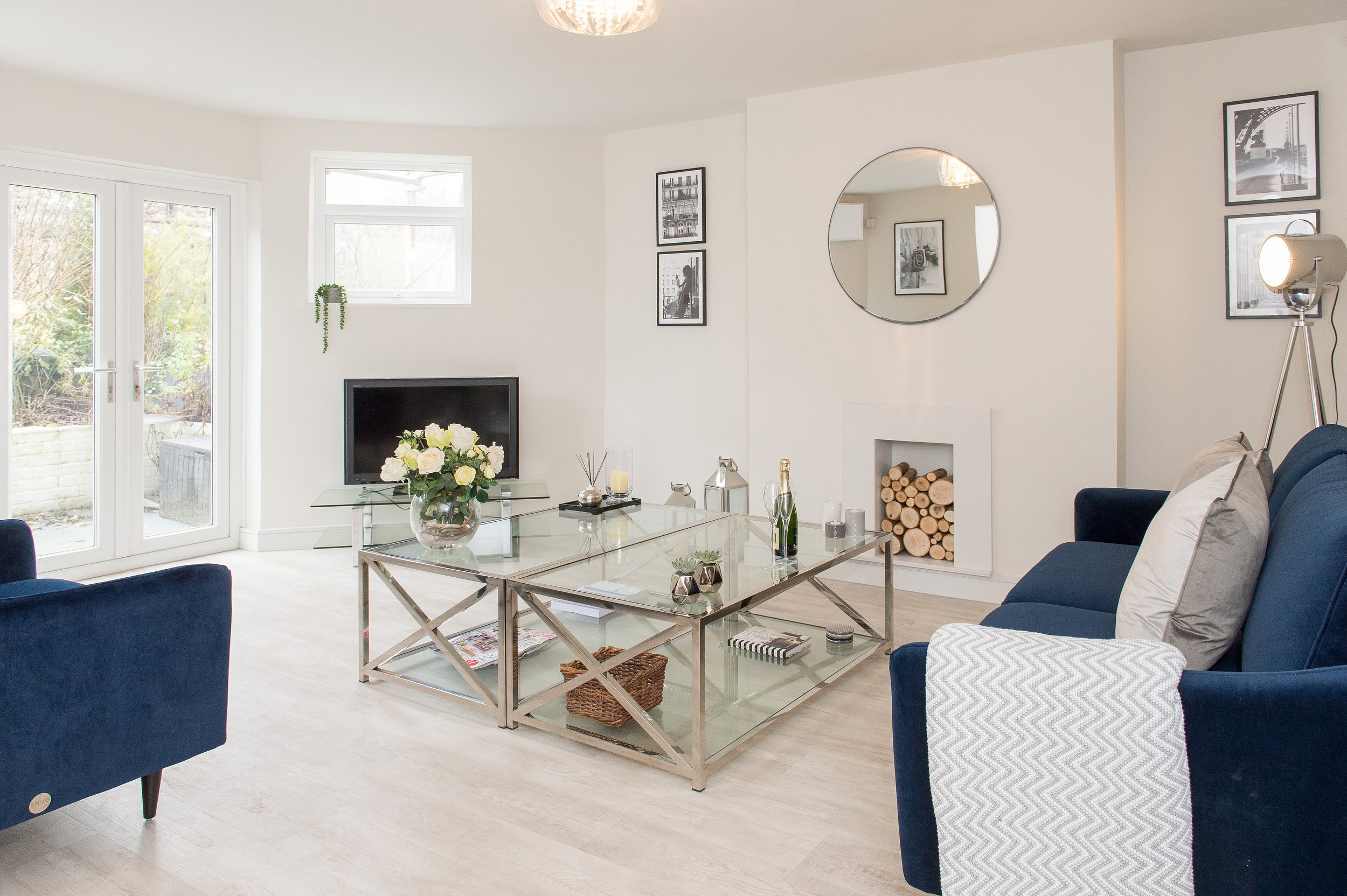 Bright living room with white walls, large windows, a glass coffee table with a flower vase, navy blue sofas, framed black-and-white art on the walls, a circular mirror, and a corner with firewood stored in a niche.