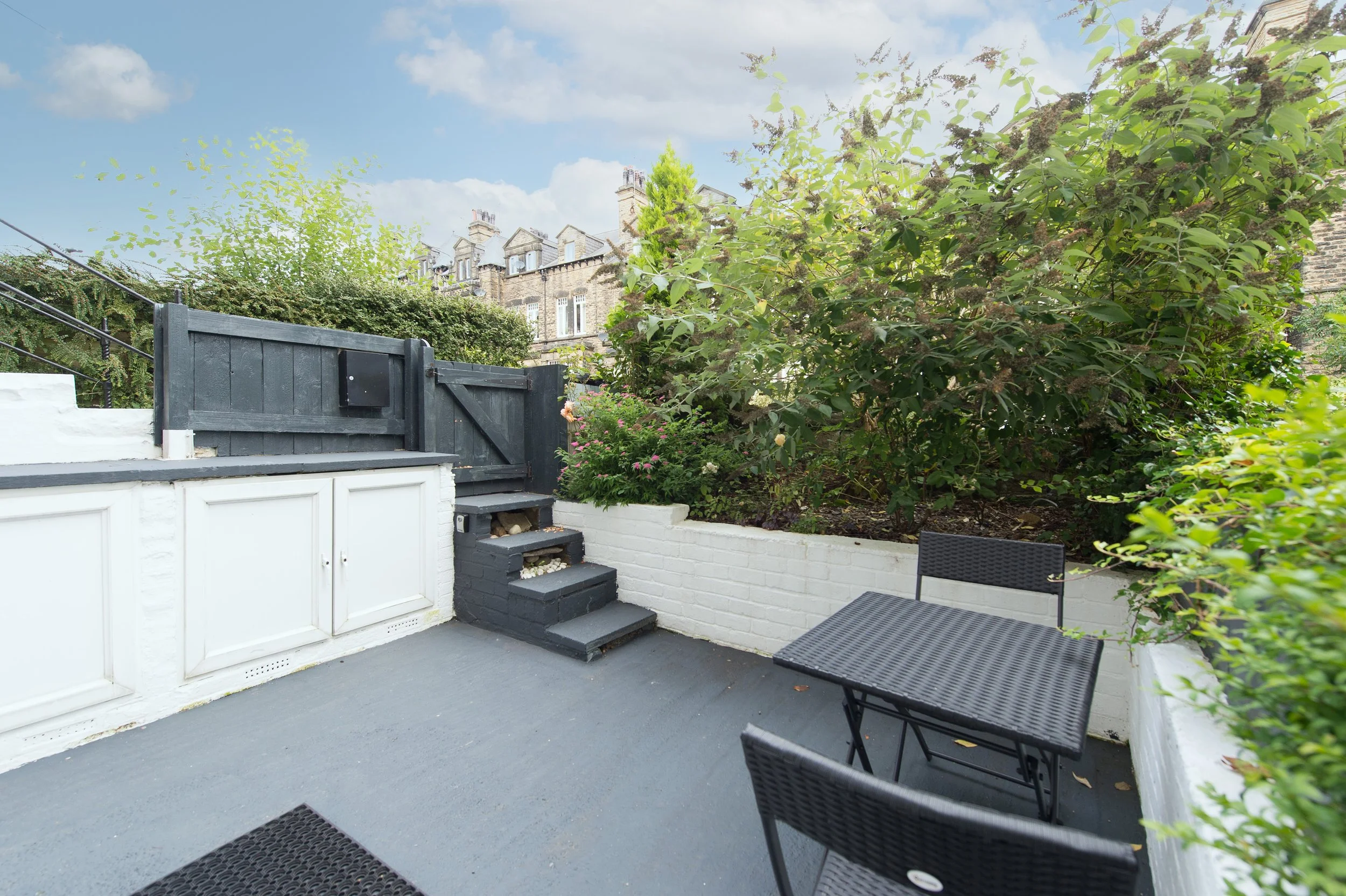 A small outdoor patio area with a grey floor, black chairs, white and black fences, greenery including bushes and small trees, and a background of old brick buildings under a cloudy sky.