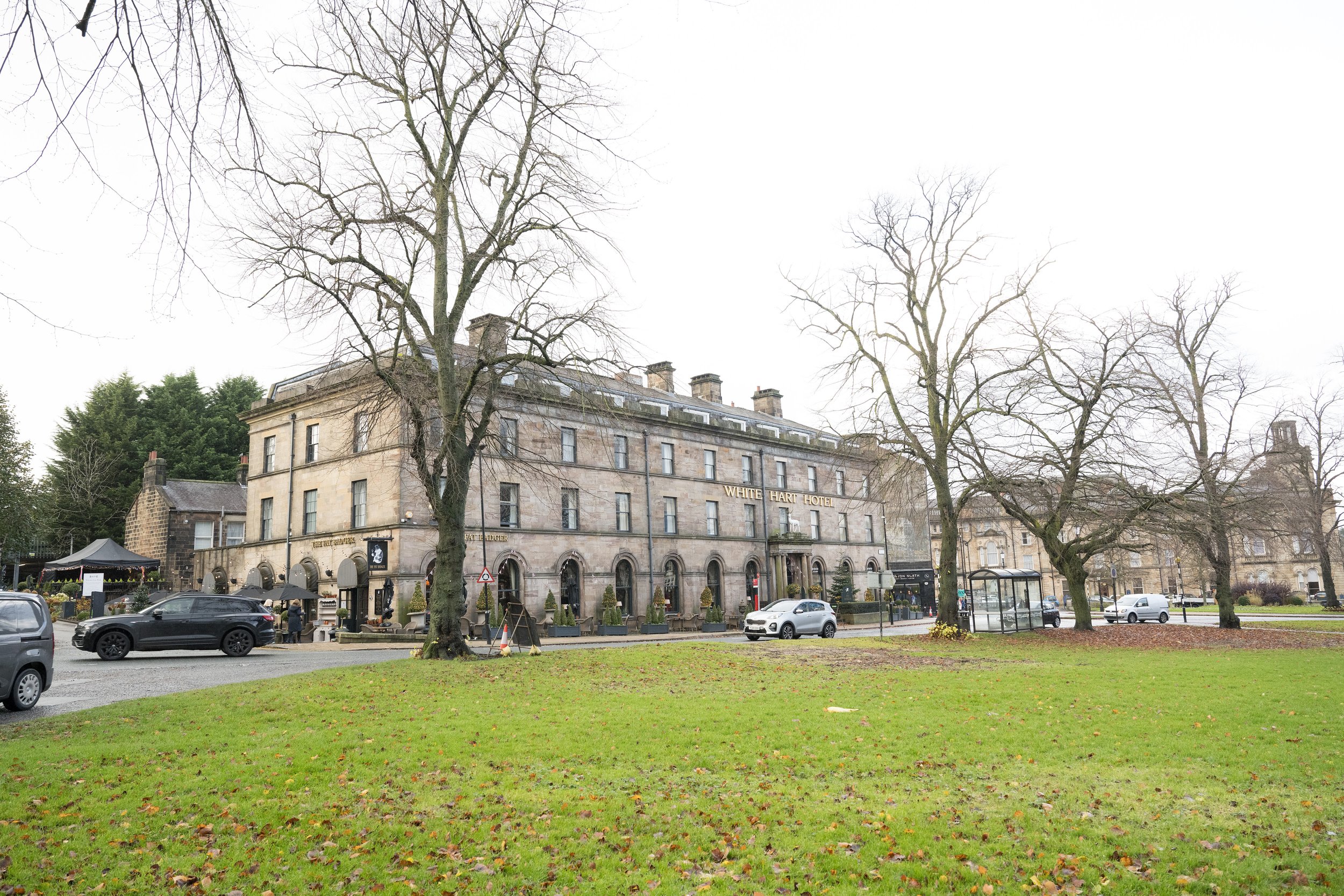 A large, historic stone hotel called the White Hart Hotel with leafless trees in front, parked cars along the street, and a cloudy sky overhead.