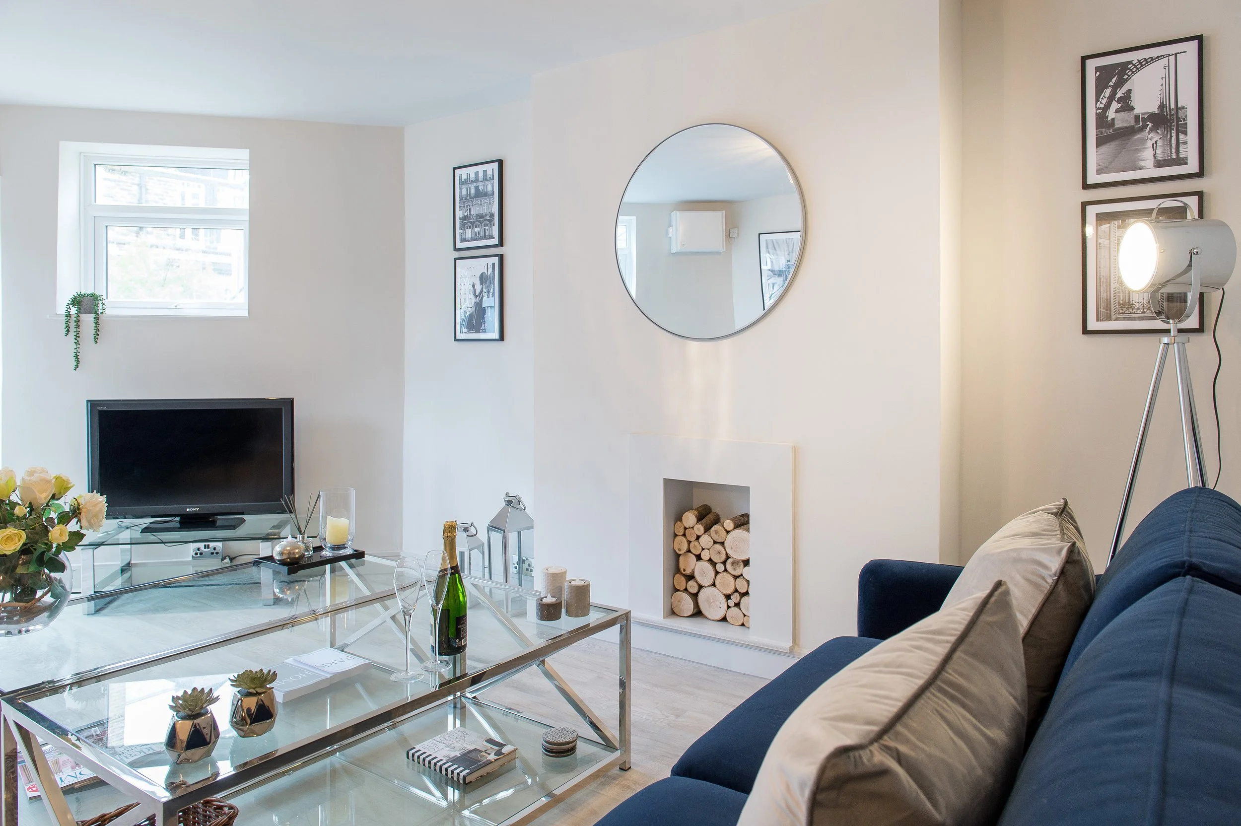 Living room with a glass coffee table, a television, framed black and white photographs on the wall, a large round mirror, a floor lamp, and a navy blue sofa with beige pillows. A small window with a potted plant below it and a wood log storage area 