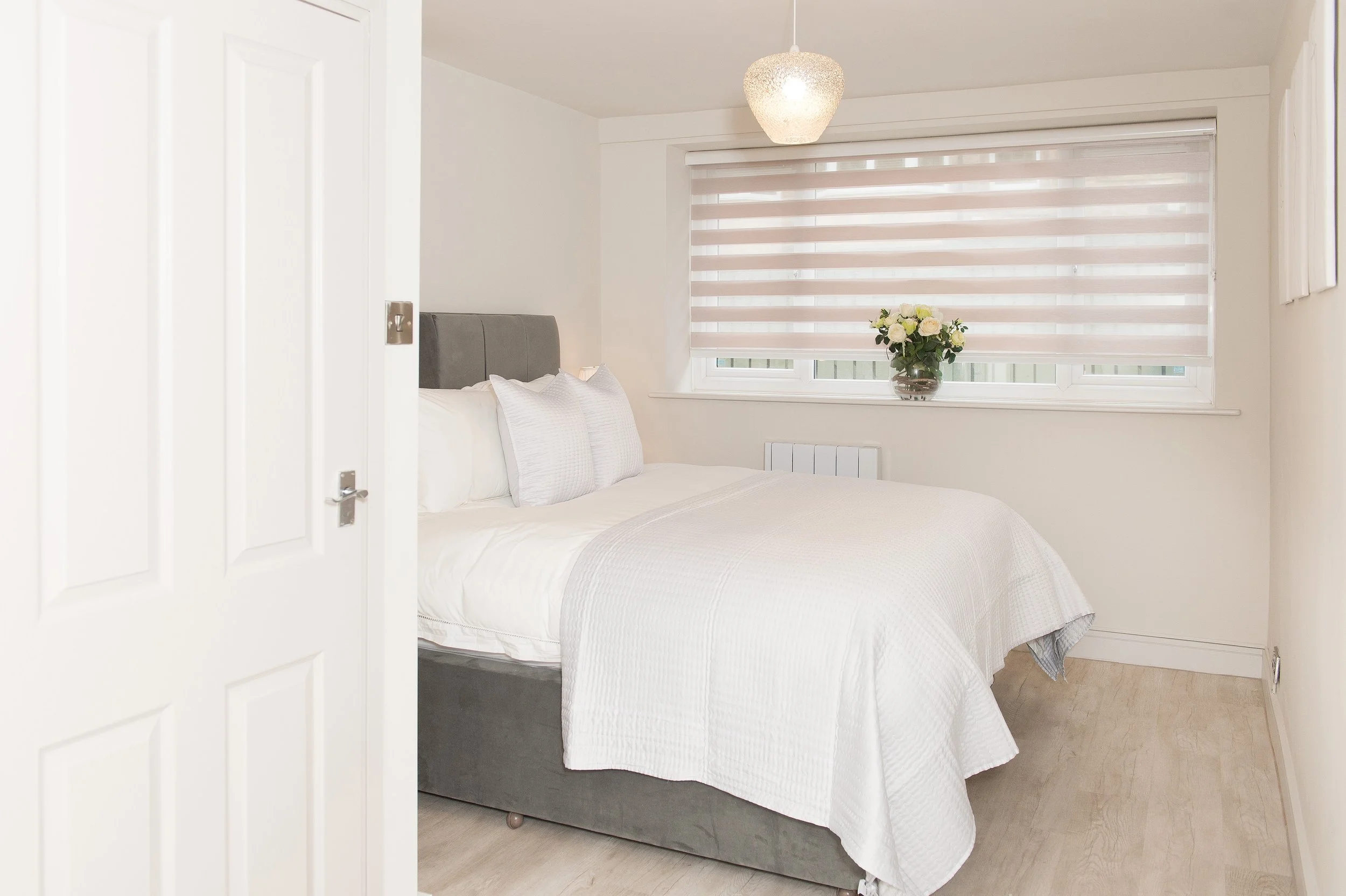 A bedroom with a bed covered in white bedding and pillows, a window with horizontal blinds, a vase with white roses, and a ceiling light fixture.
