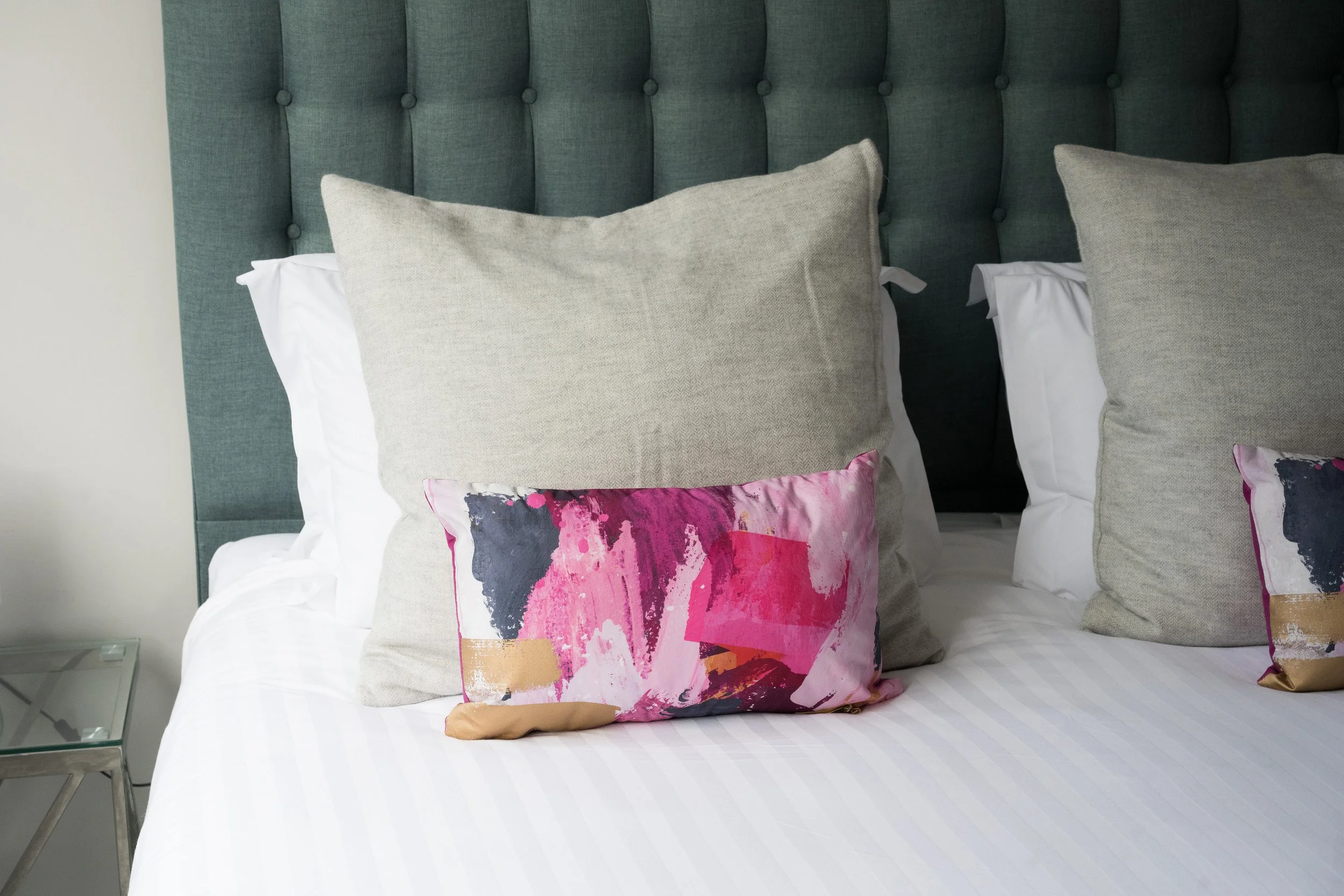 Close-up of a bed with a light gray upholstered headboard, adorned with multiple decorative pillows including two large plain gray pillows, and three smaller abstract patterned pillows featuring pink, black, and beige colors.