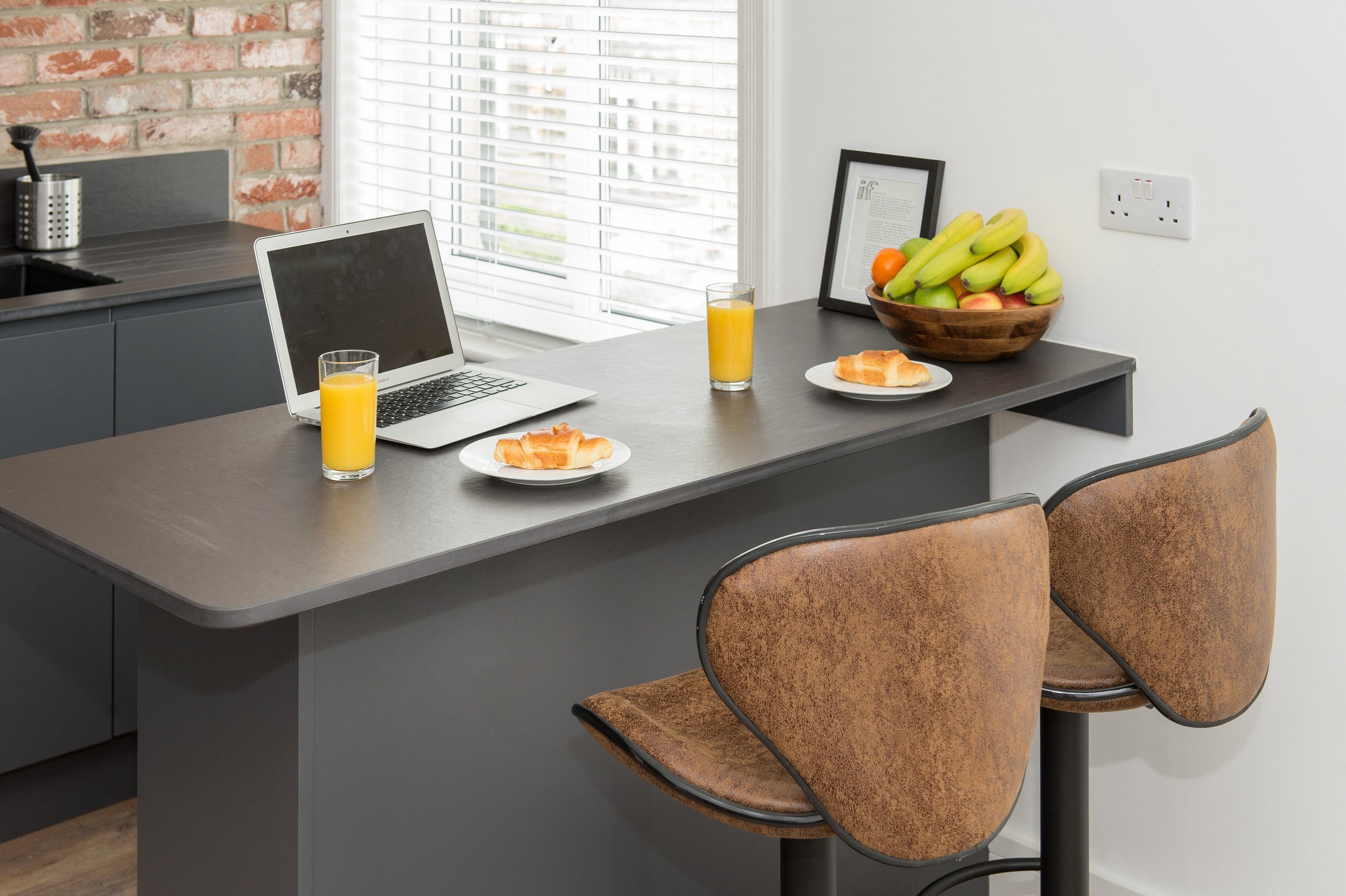 A kitchen with a gray counter and a brick wall. There is a laptop, two glasses of orange juice, two plates with croissants, a framed picture, a bowl of bananas and some fruit on the counter. A window with blinds is in the background.