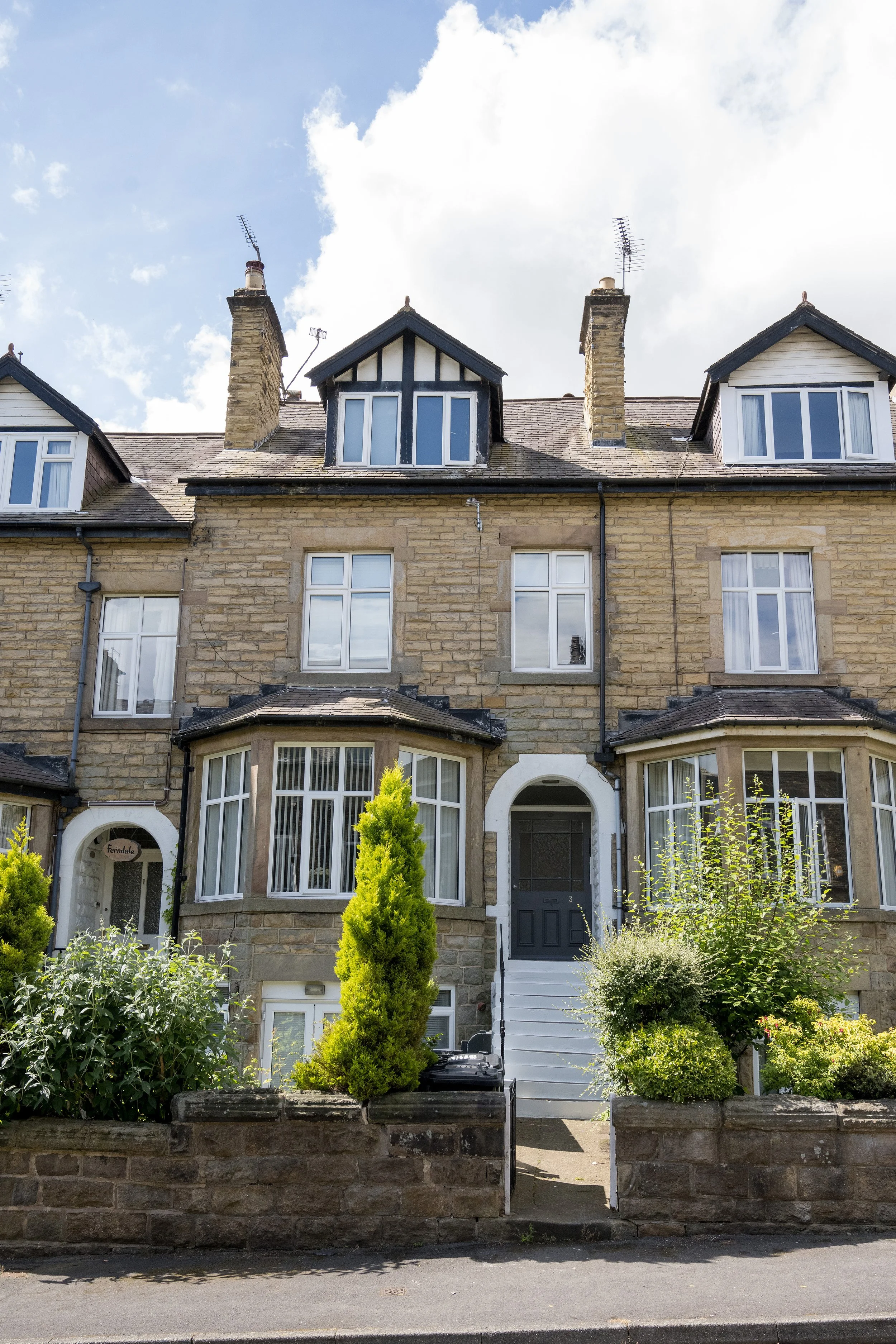 A multi-story stone and brick house with pointed gabled roofs, white-framed windows, and a small front garden with bushes and trees. The house has three large attic windows with dormer roofs, and there are brick chimneys with TV antennas.