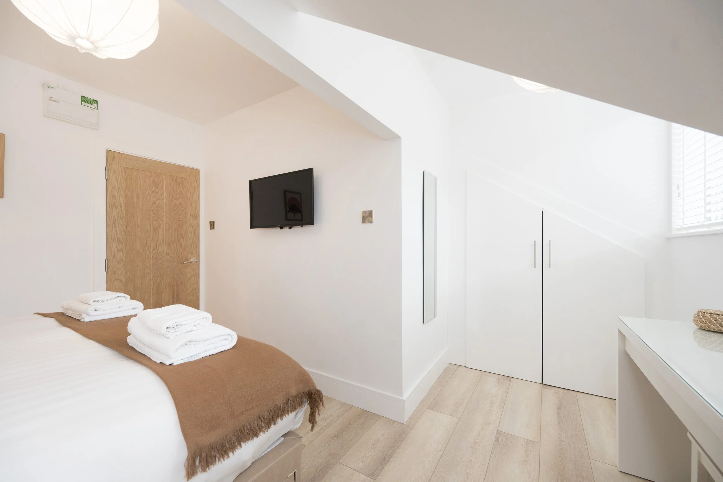 A minimalistic white bedroom with a neatly made bed, folded towels on the bed, a wooden door, a wall-mounted flat-screen TV, a white built-in wardrobe, and a window with blinds letting in natural light.