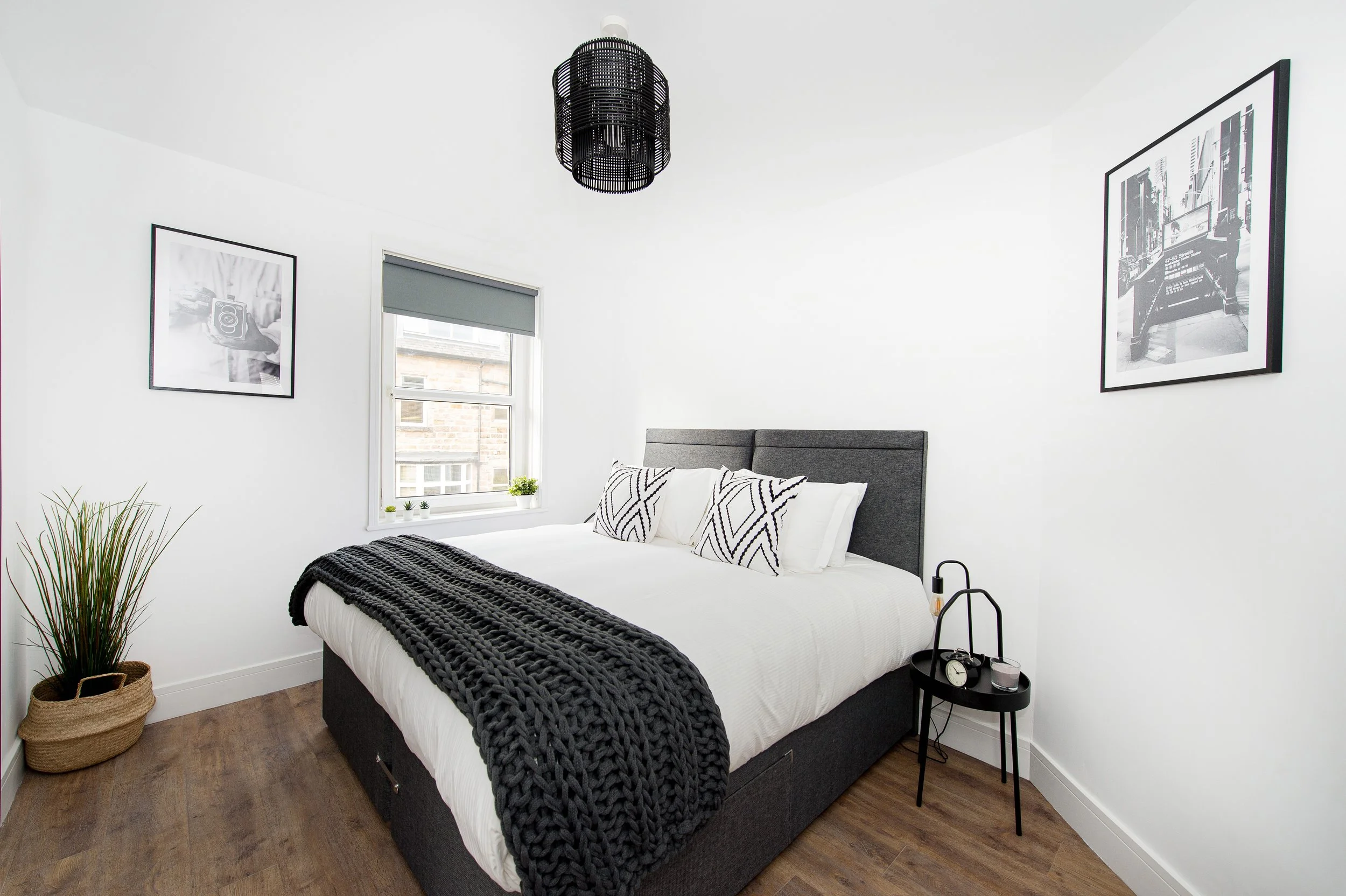 A modern minimalist bedroom with a large bed with white bedding, black and white patterned pillows, and a chunky black knitted blanket. There is a black bedside table with a clock, candles, and a glass on it. A black decorative pendant light hangs fr