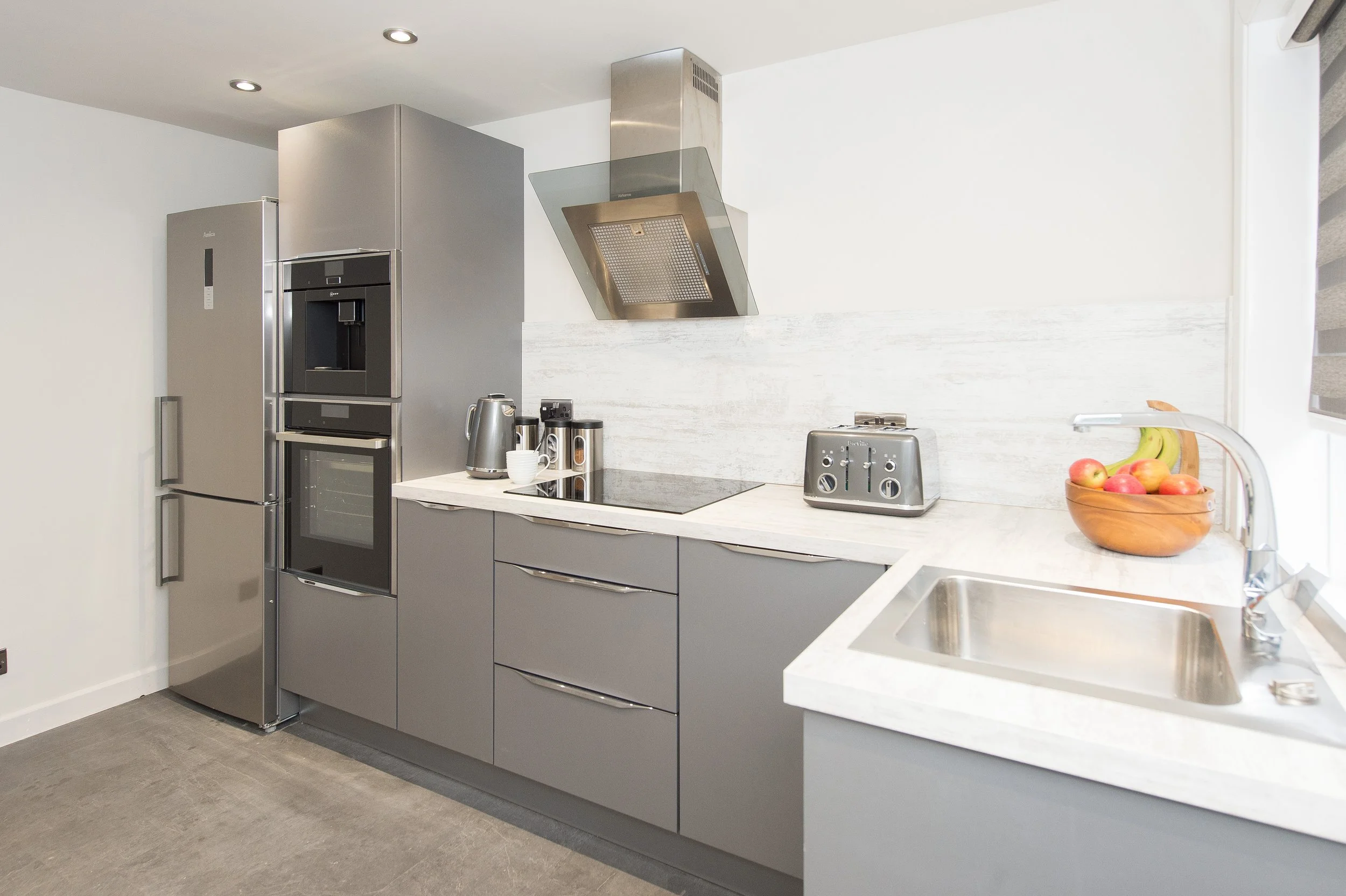 Modern kitchen with gray cabinets, white marble countertops, stainless steel refrigerator, oven, microwave, and range hood. A bowl of apples is on the counter near the sink.