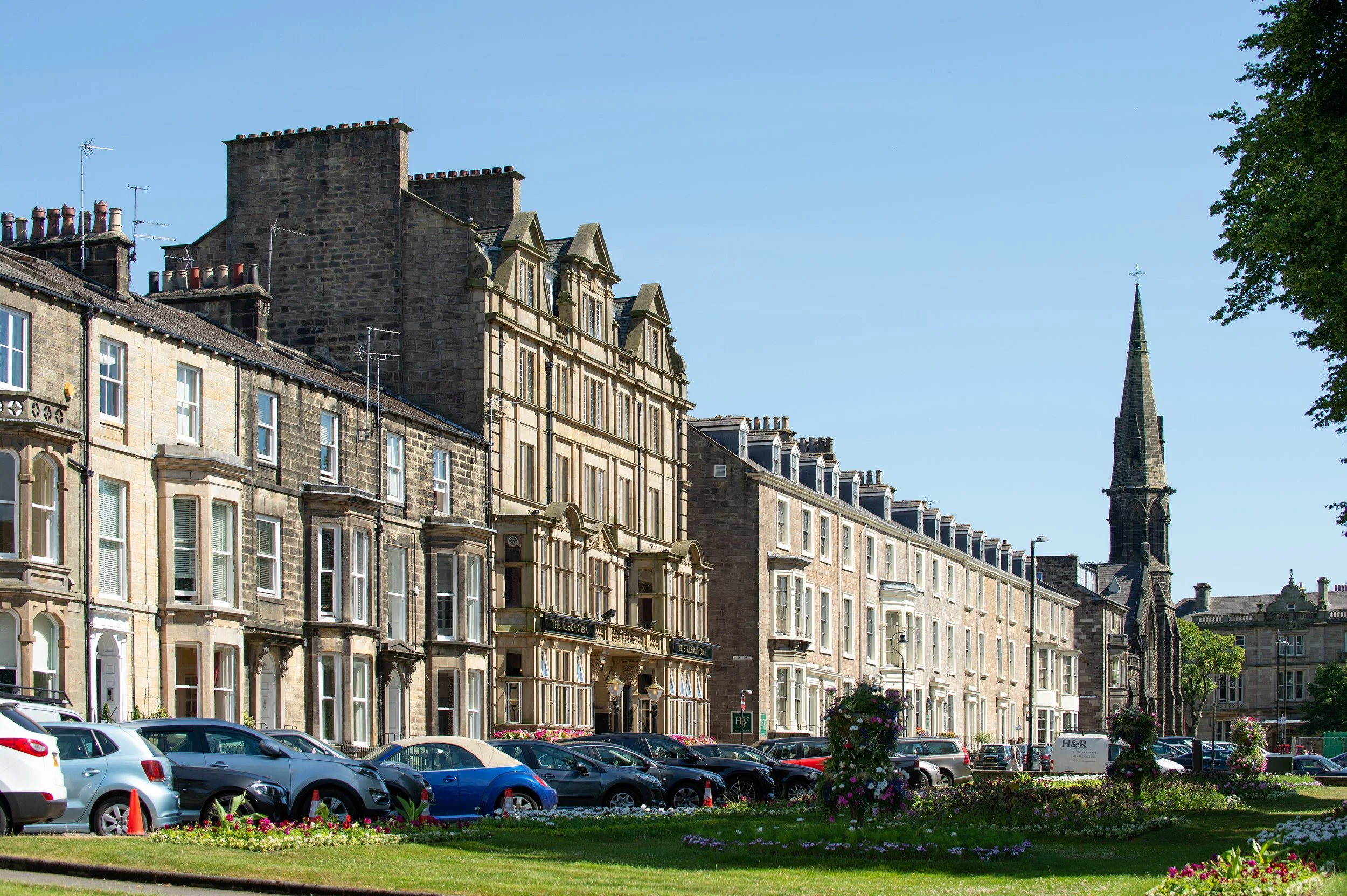 A row of historic multi-story buildings with ornate architecture along a street, with a parking lot filled with cars and a grassy area with flowers in the foreground. A church steeple is visible in the background against a clear blue sky.