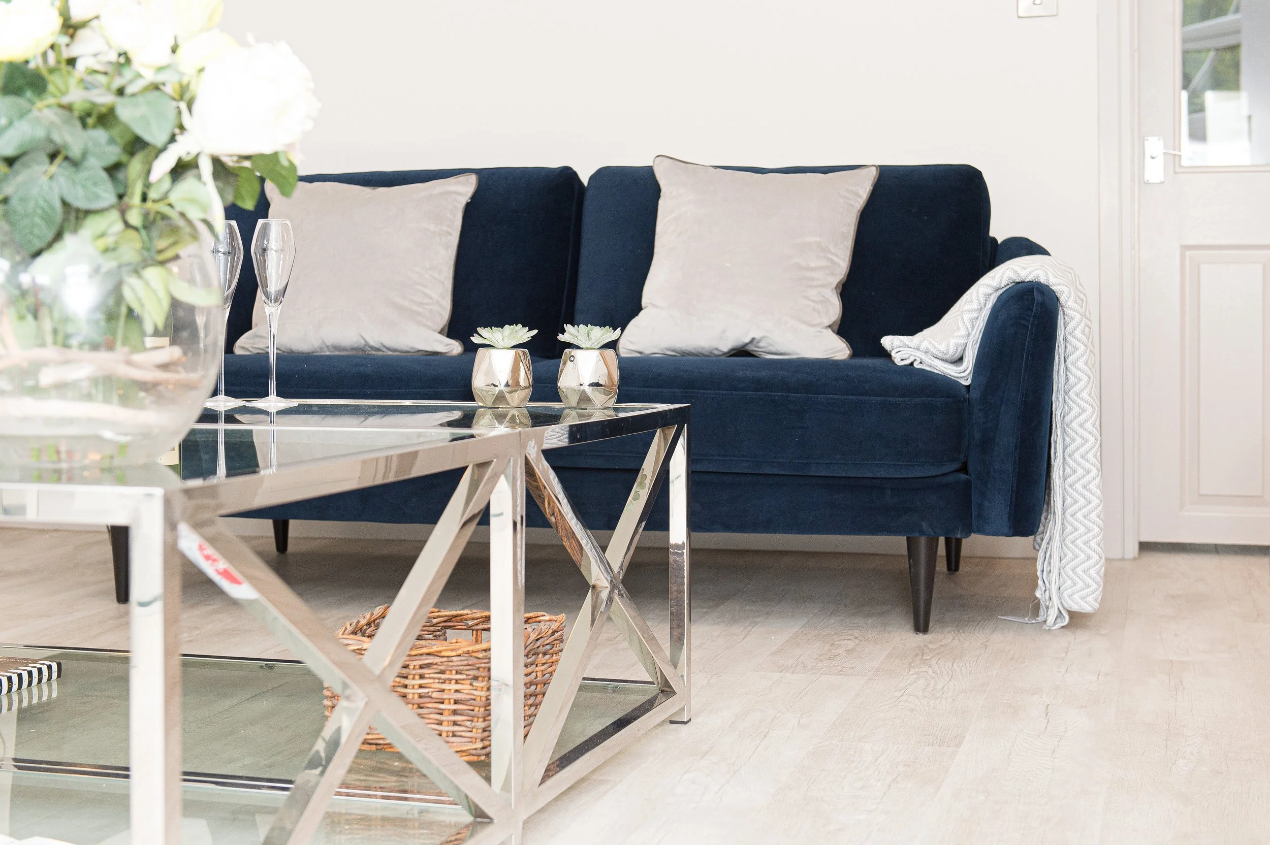 Living room with a navy blue sofa, light-colored pillows, glass coffee table, and decorative plants.