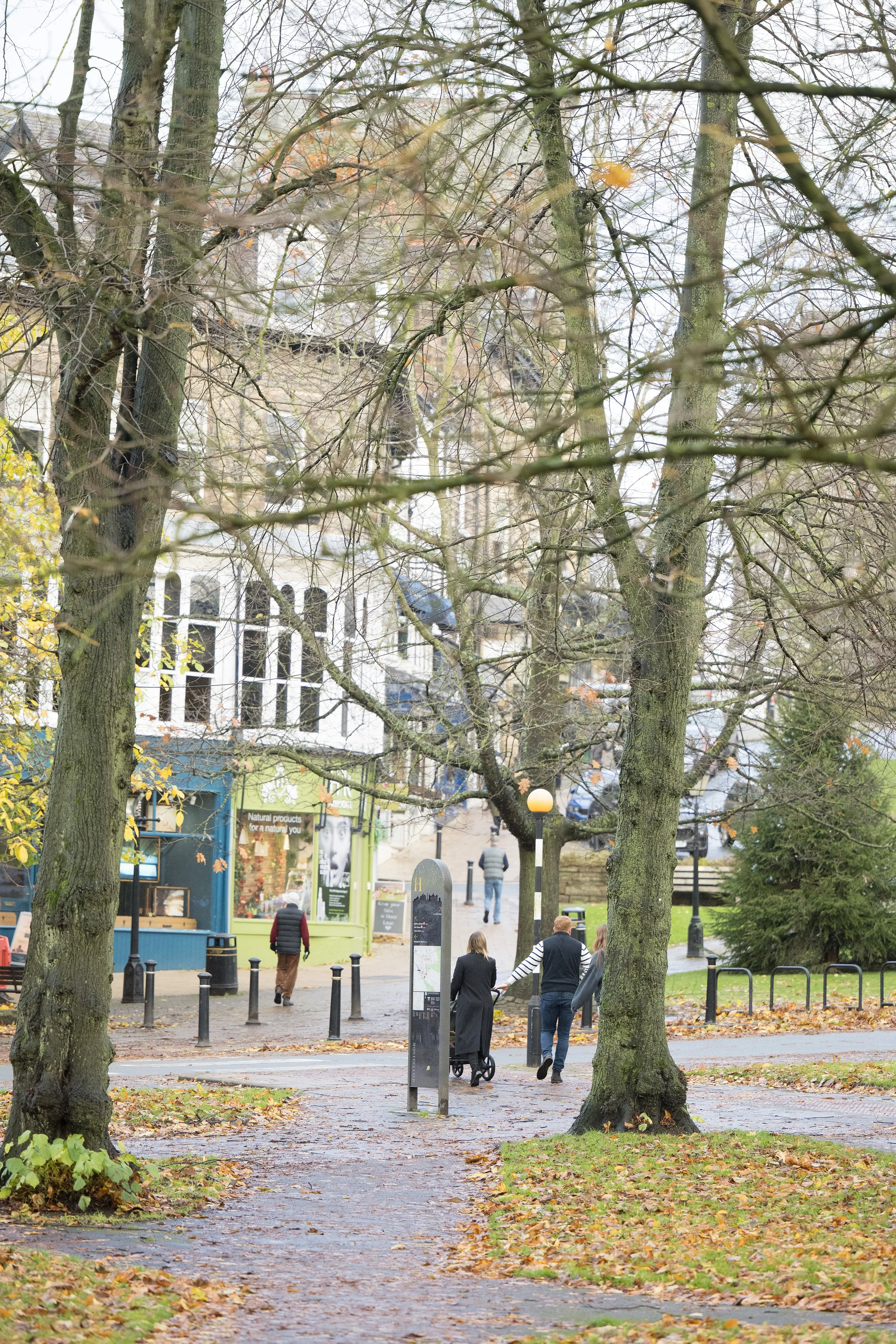 A park scene with pedestrians walking along a paved pathway lined with trees shedding yellow and brown leaves, with small shops in the background and street lamps.