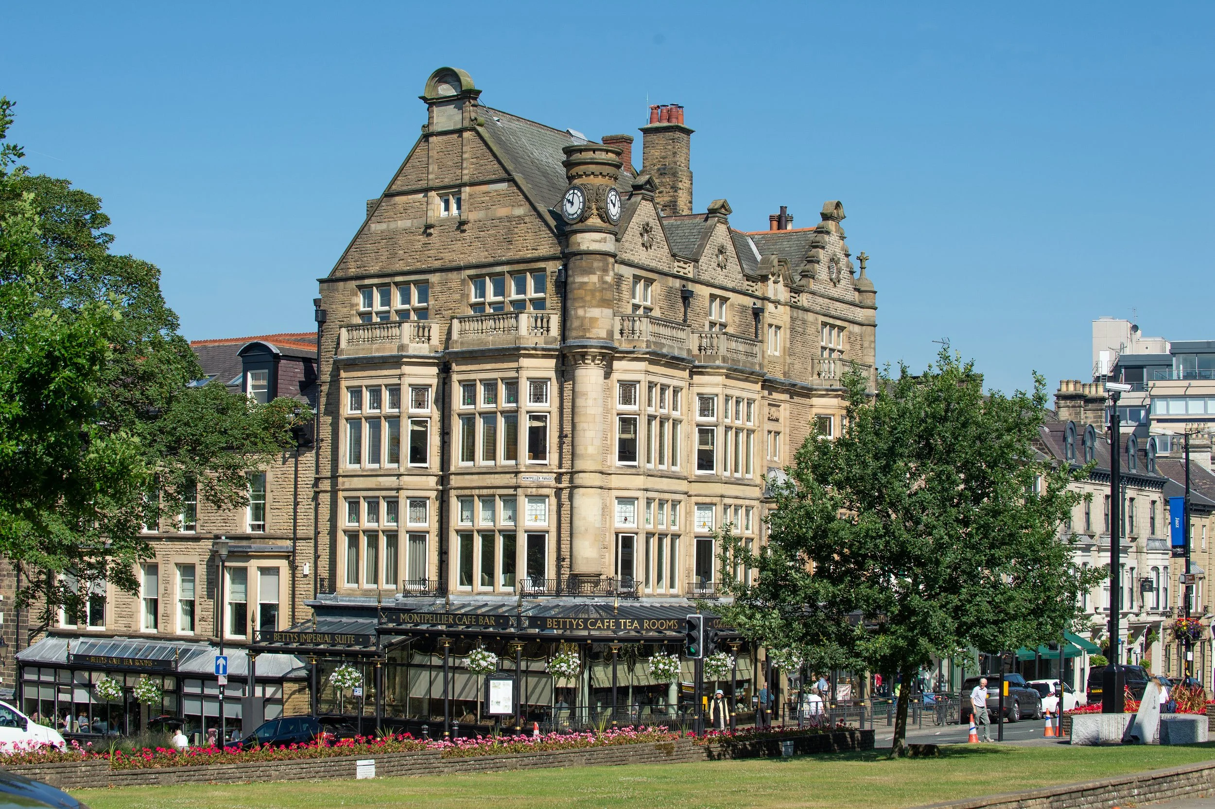 A historic building with a clock tower, large windows, and ornate stone architecture. The building houses Bettys Cafe Tea Rooms, with visible signage. In the foreground, there is a park with flowers, trees, and people walking, along with cars parked 