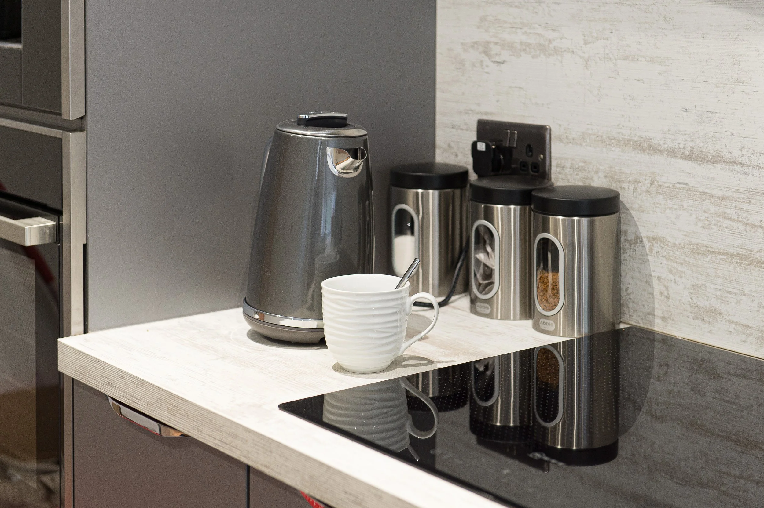 Kitchen counter with a coffee grinder, a ceramic mug with a spoon, and three stainless steel containers with black lids, plugged into an electrical outlet.