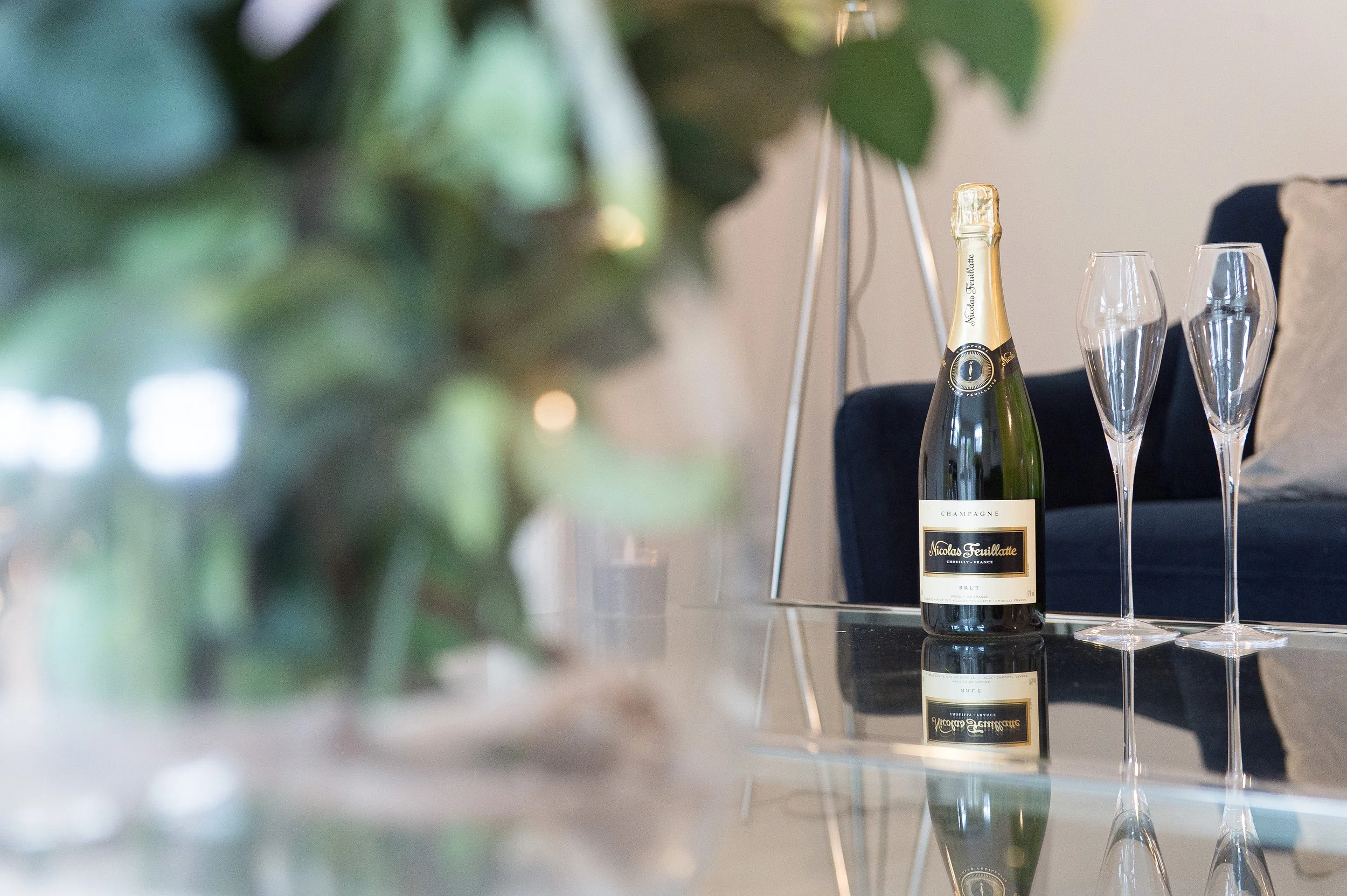 A bottle of Nicolas Feuillatte champagne and two empty champagne glasses on a reflective surface in a living room setting.