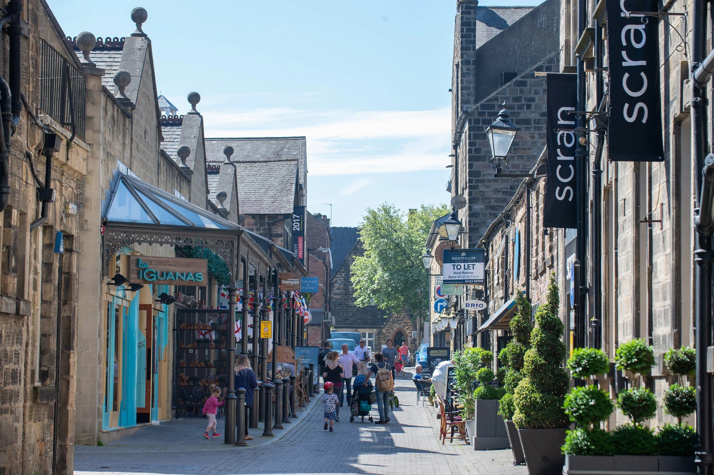 A bustling pedestrian street lined with shops and cafes, with people walking, shopping, and pushing strollers under a bright blue sky.