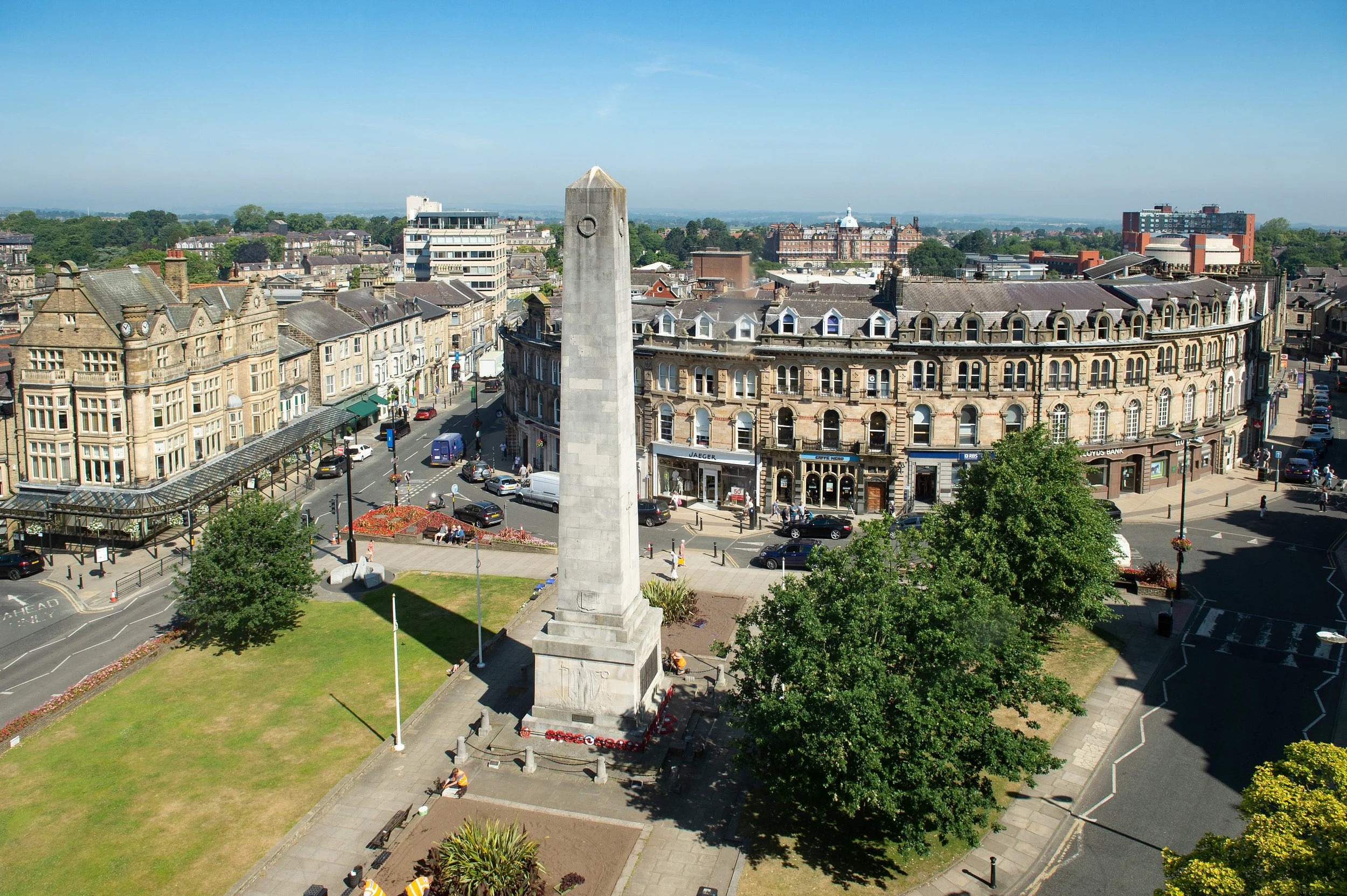 A panoramic view of a historic city street with classic stone buildings, a central monument, and modern high-rise structures under a clear blue sky.
