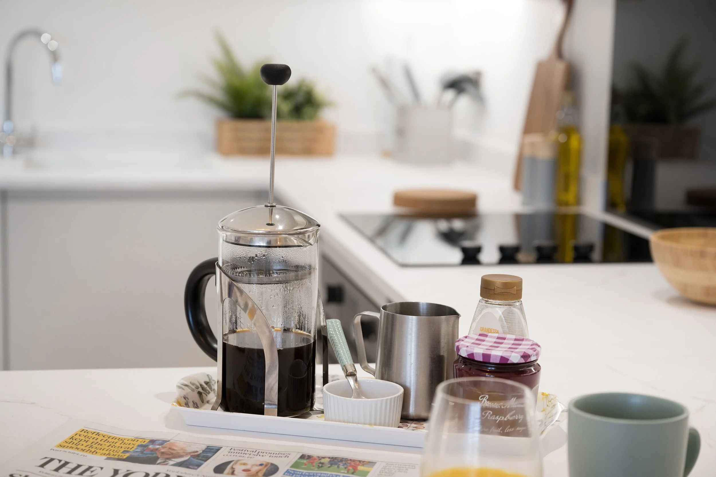 Kitchen scene with French press coffee maker, mugs, and jars on a white countertop. In the background are kitchen utensils, plants, and bottles, with a newspaper on the table in the foreground.