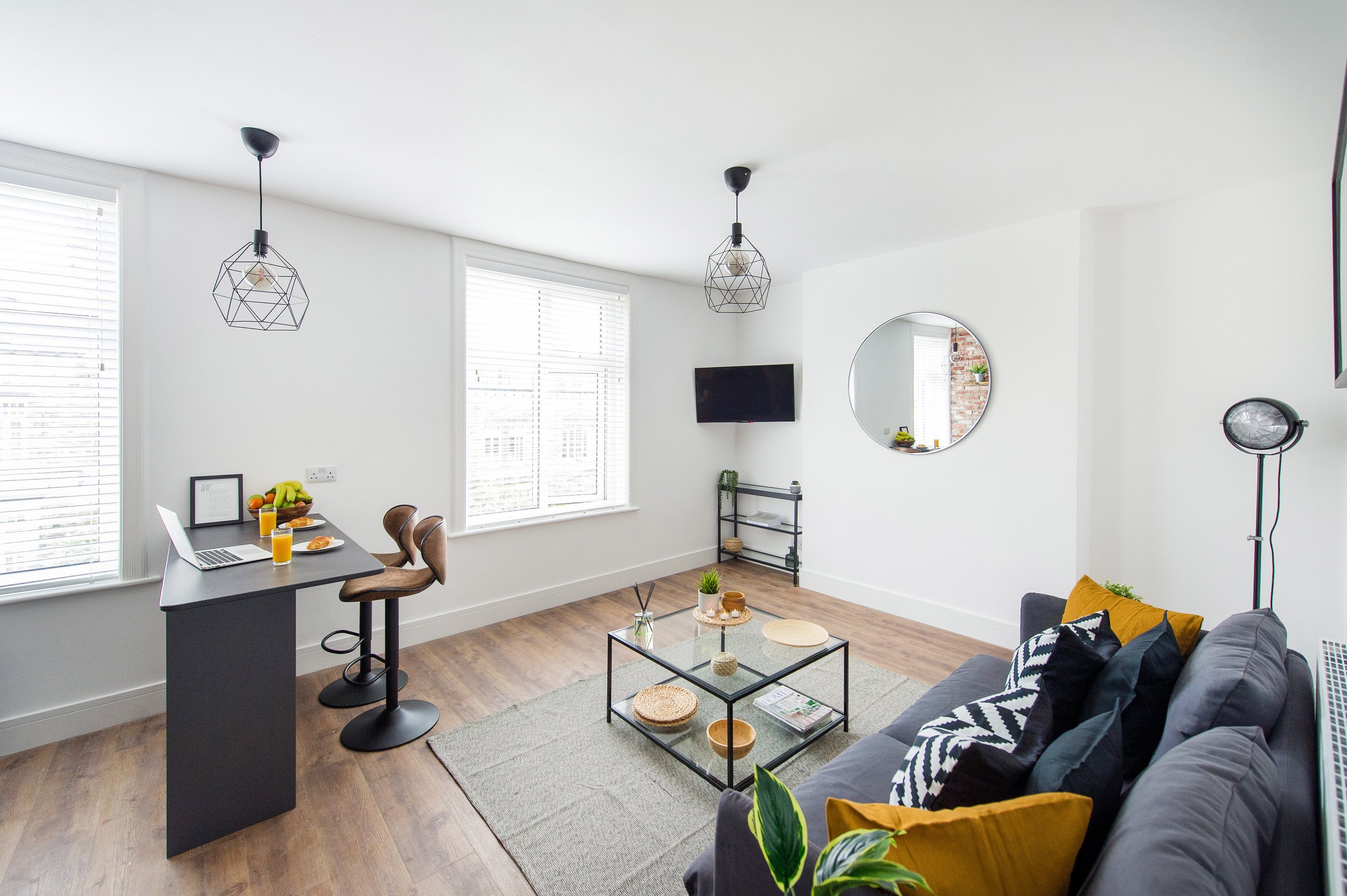 Modern living room with large windows, a gray sofa with black and white pillows, glass coffee table, TV mounted on white wall, round mirror, black metal shelf, and two geometric pendant lights.