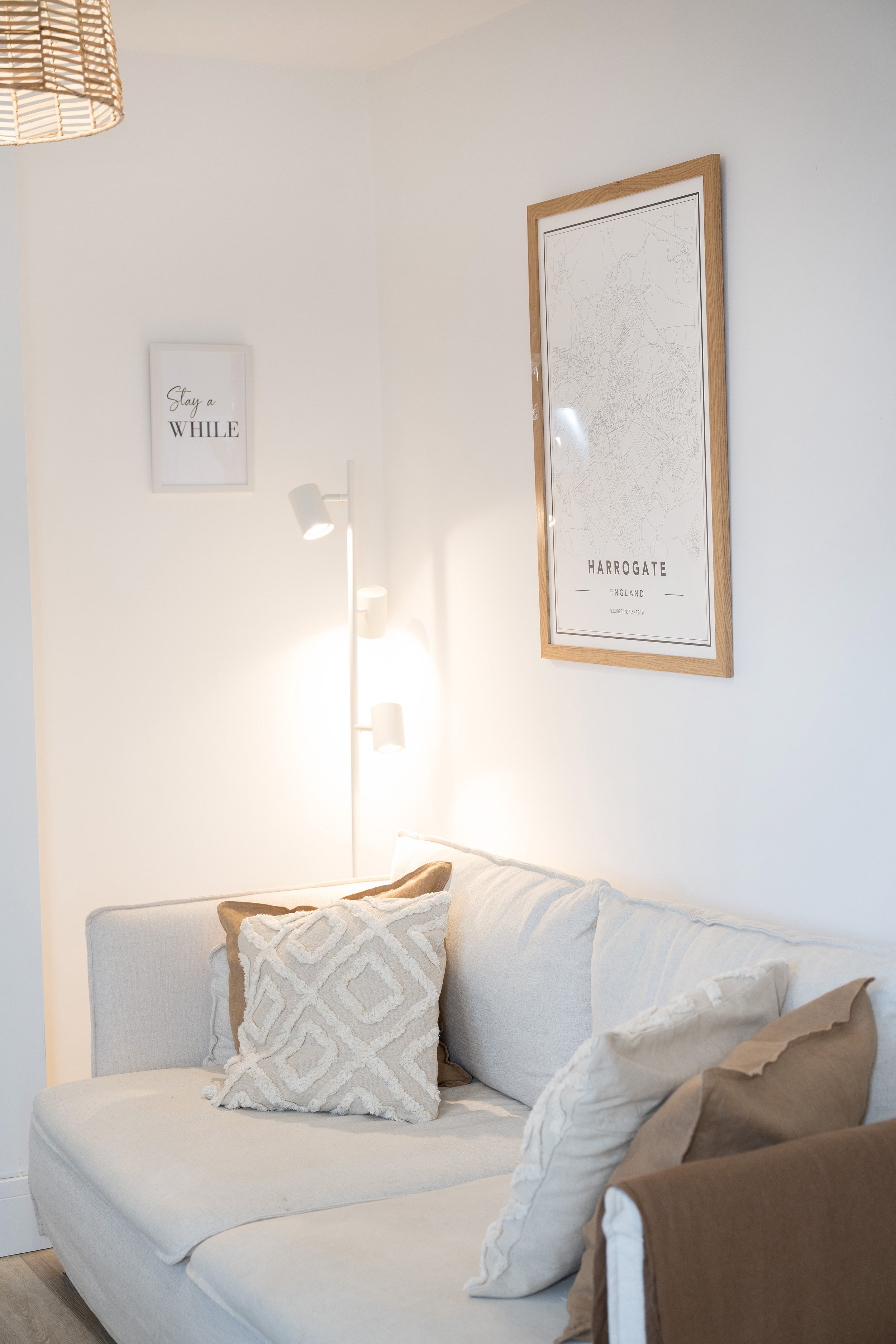 A cozy living room with a beige sofa filled with decorative pillows, a white wall with framed art featuring a map of Harrogate, England, and a white floor lamp with two adjustable light fixtures.