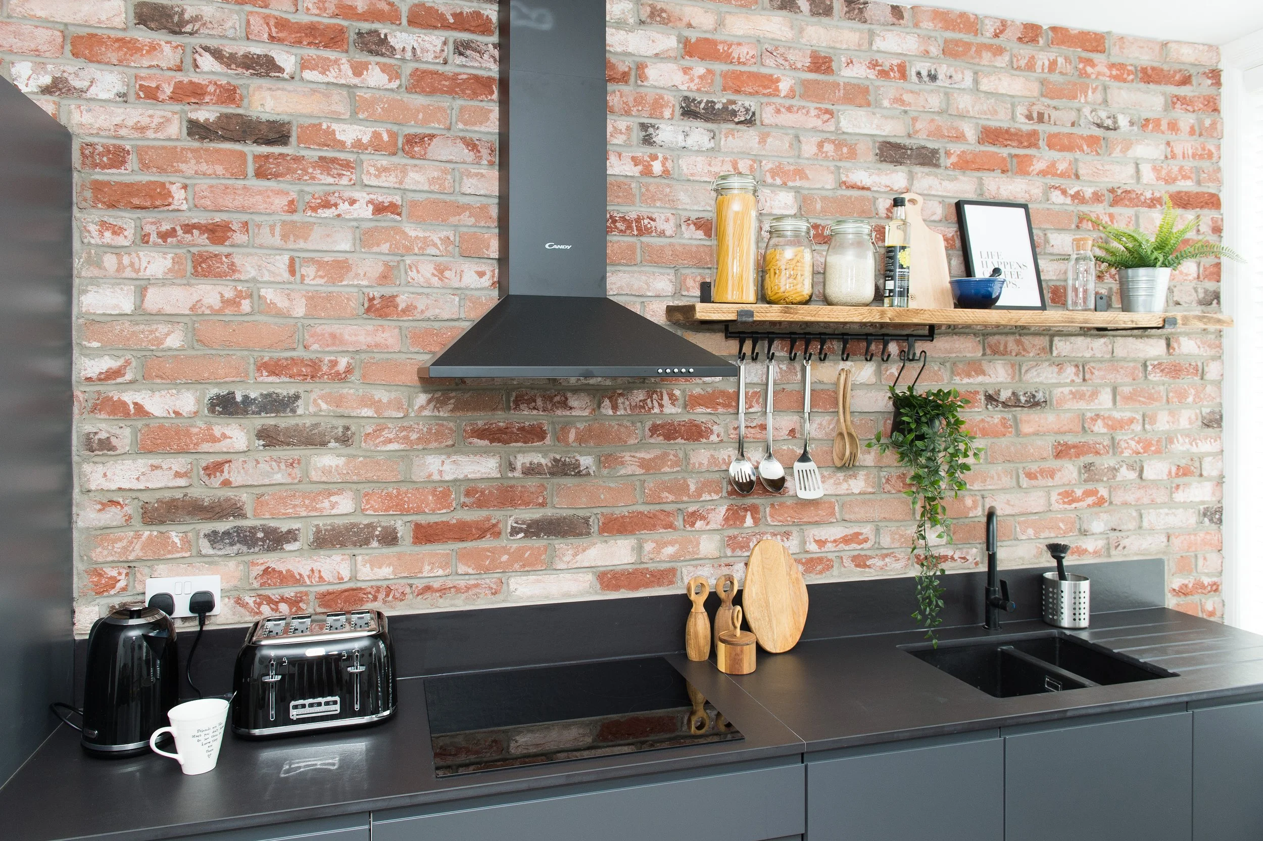 Modern kitchen with exposed brick wall, black countertop, and shelving holding jars of pasta, a framed quote, a potted fern, and kitchen utensils. Counter has a coffee machine, toaster, and a mug.
