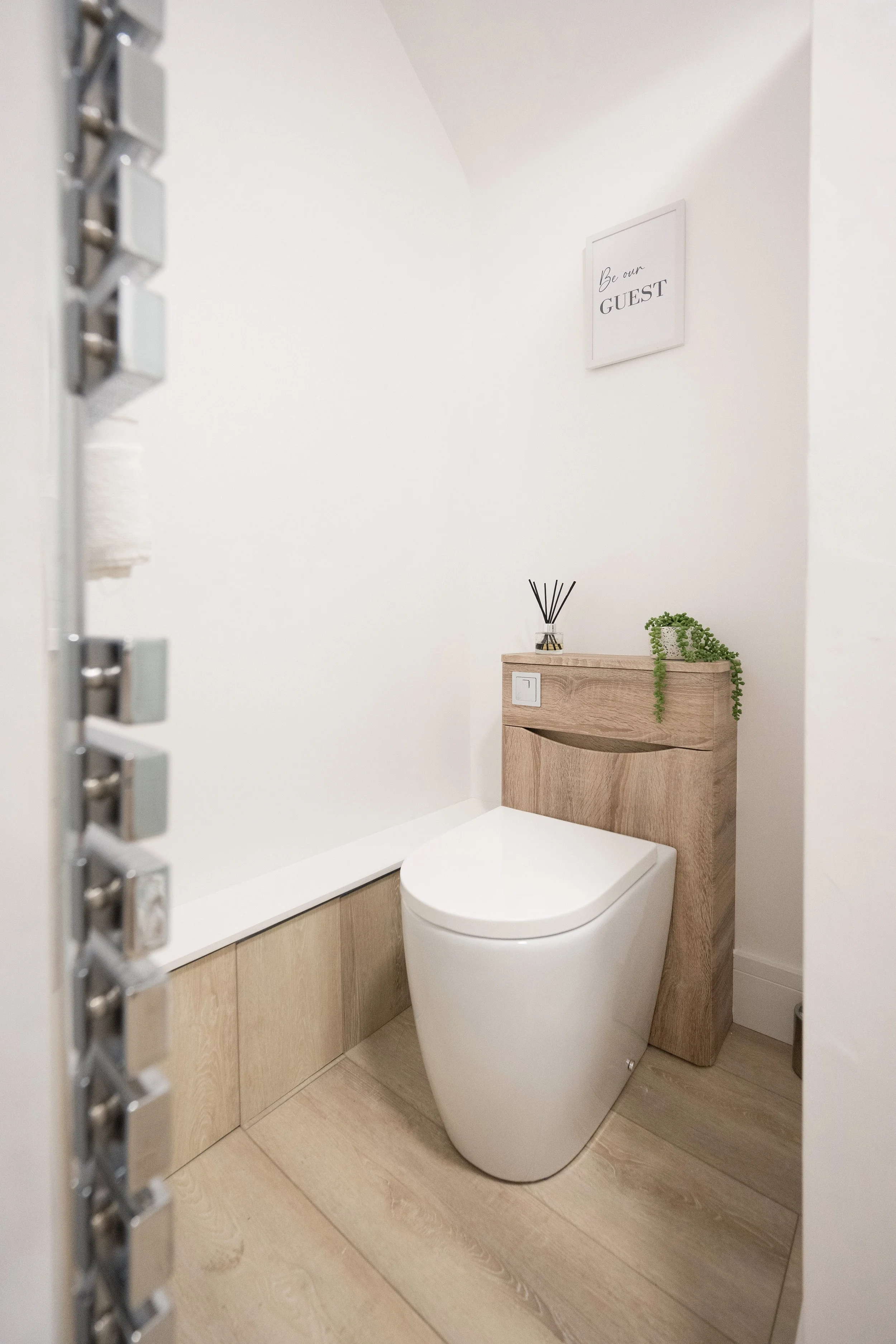 A modern small bathroom with a white toilet, light wood cabinetry, a wall decoration reading 'Be our GUEST', and decorative objects like a reed diffuser and a trailing green plant on the cabinet.