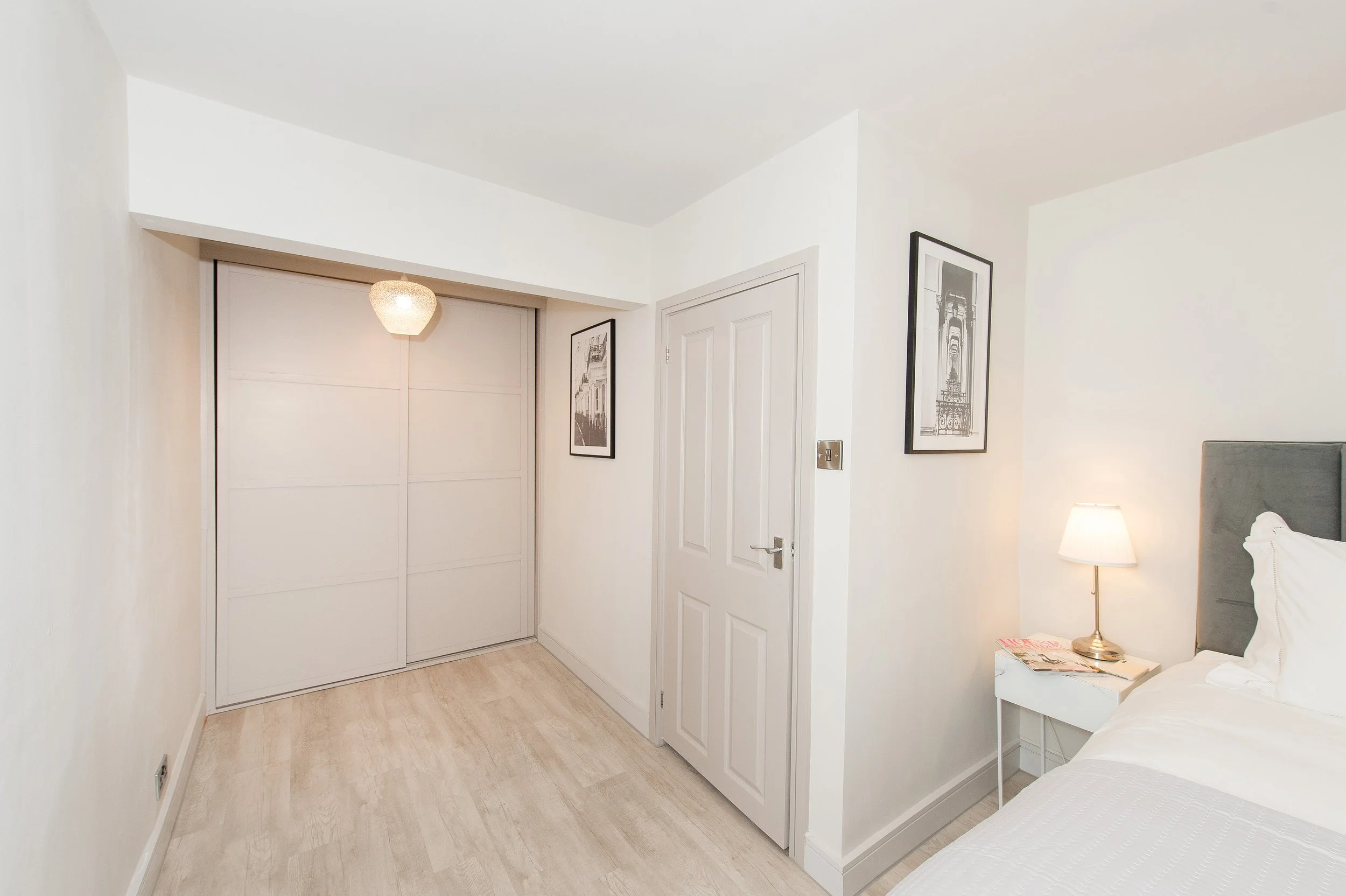 A minimalist bedroom with white walls, a bed with white bedding, a nightstand with a lamp and magazines, framed black and white photos on the wall, a closed white door, and a built-in wardrobe with sliding doors.