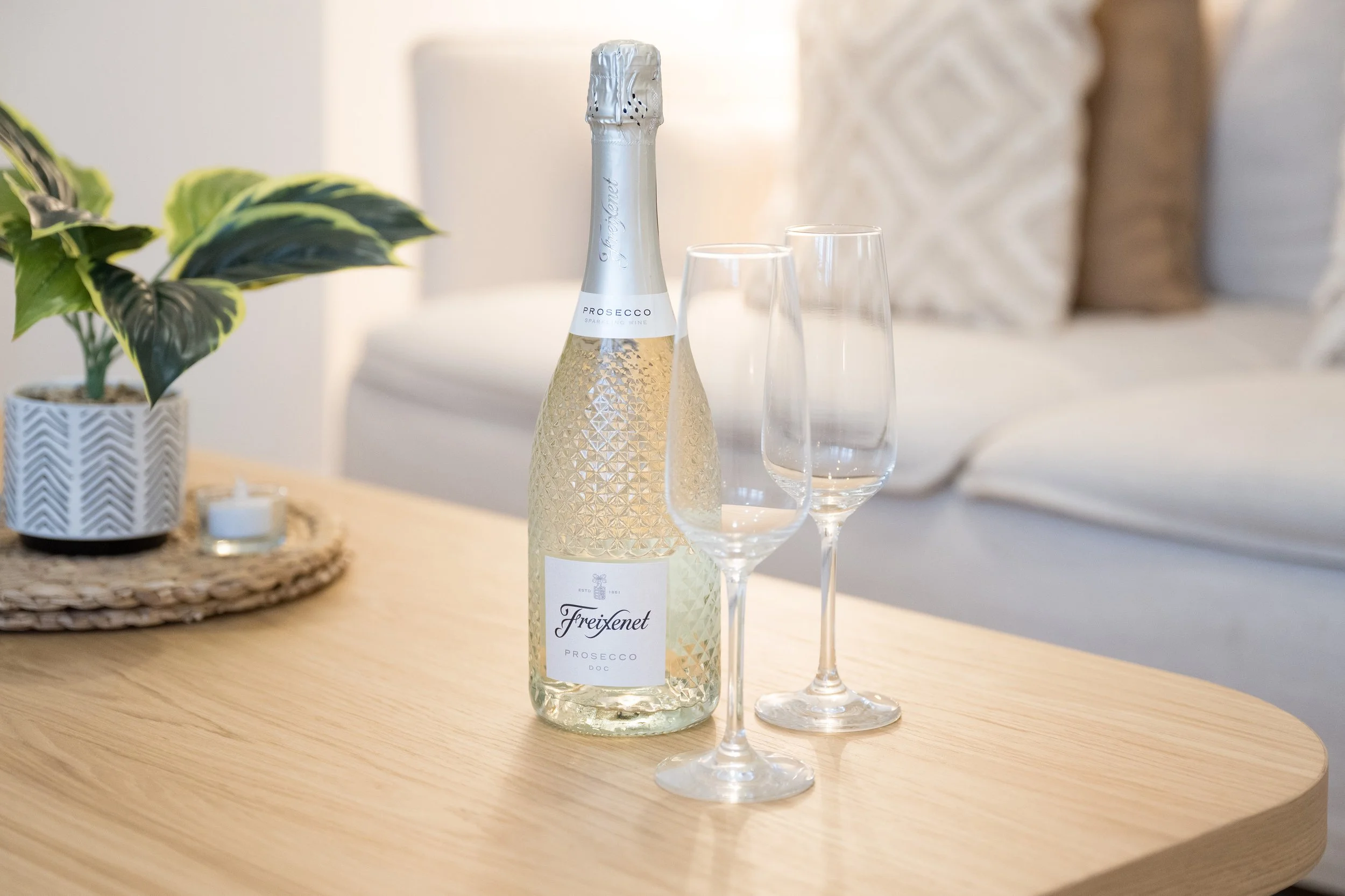 A bottle of Freixenet prosecco with a foil seal, two empty champagne flutes, a potted plant, and a small candle on a woven placemat on a wooden table in a cozy room.